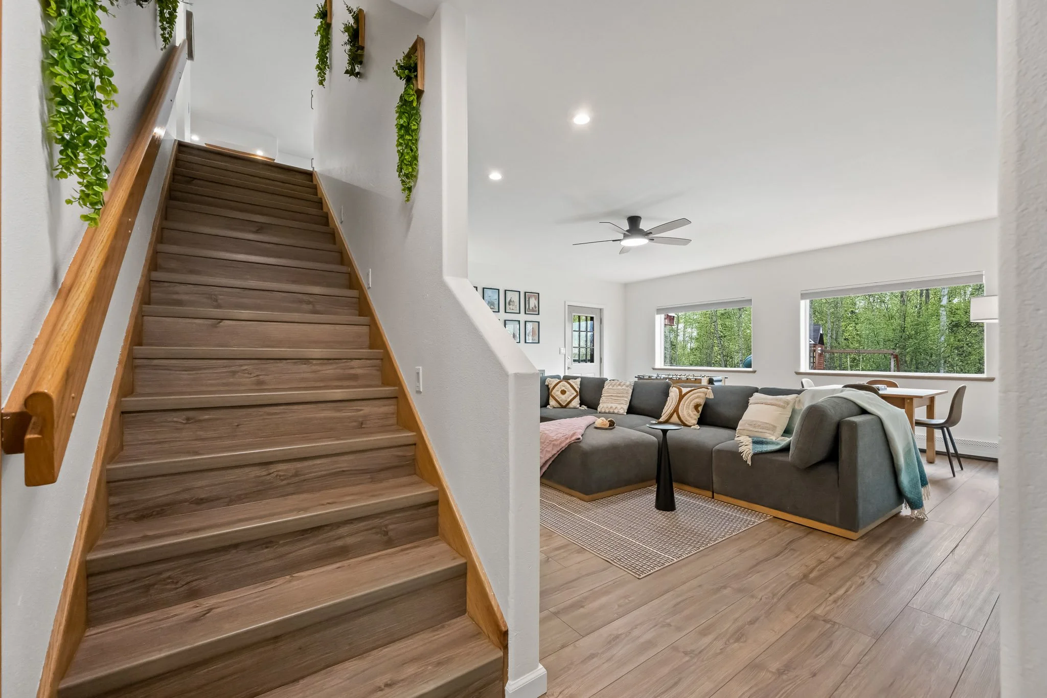 Living room with hardwood floors, large windows overlooking greenery, a gray sectional sofa with throw pillows, a black ceiling fan, and a staircase with wooden steps and a wooden handrail, decorated with hanging green plants.