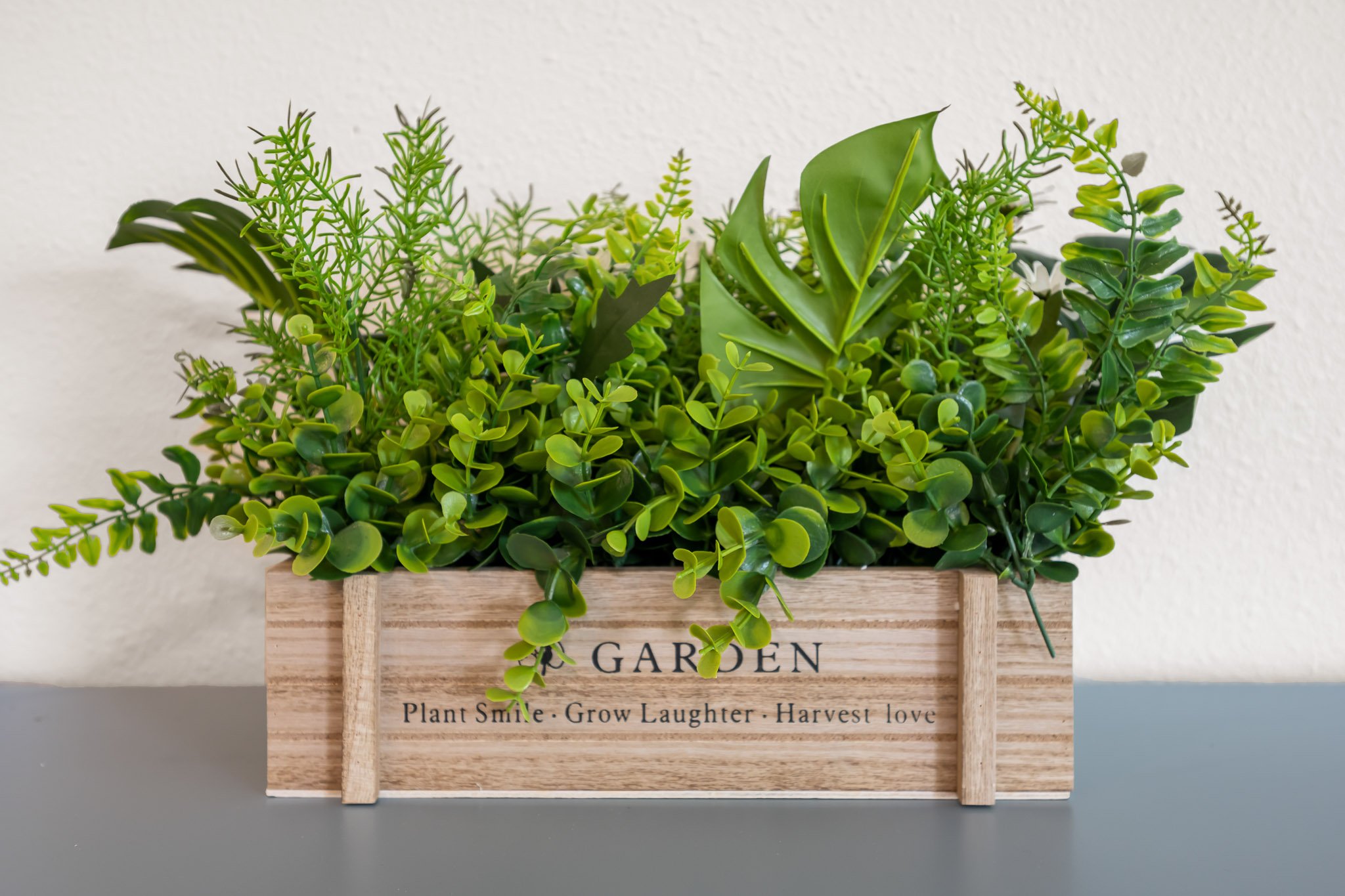 Artificial green plants in a wooden planter box with a sign that reads "GARDEN" and words below: "Plant Smile - Grow Laughter - Harvest love."