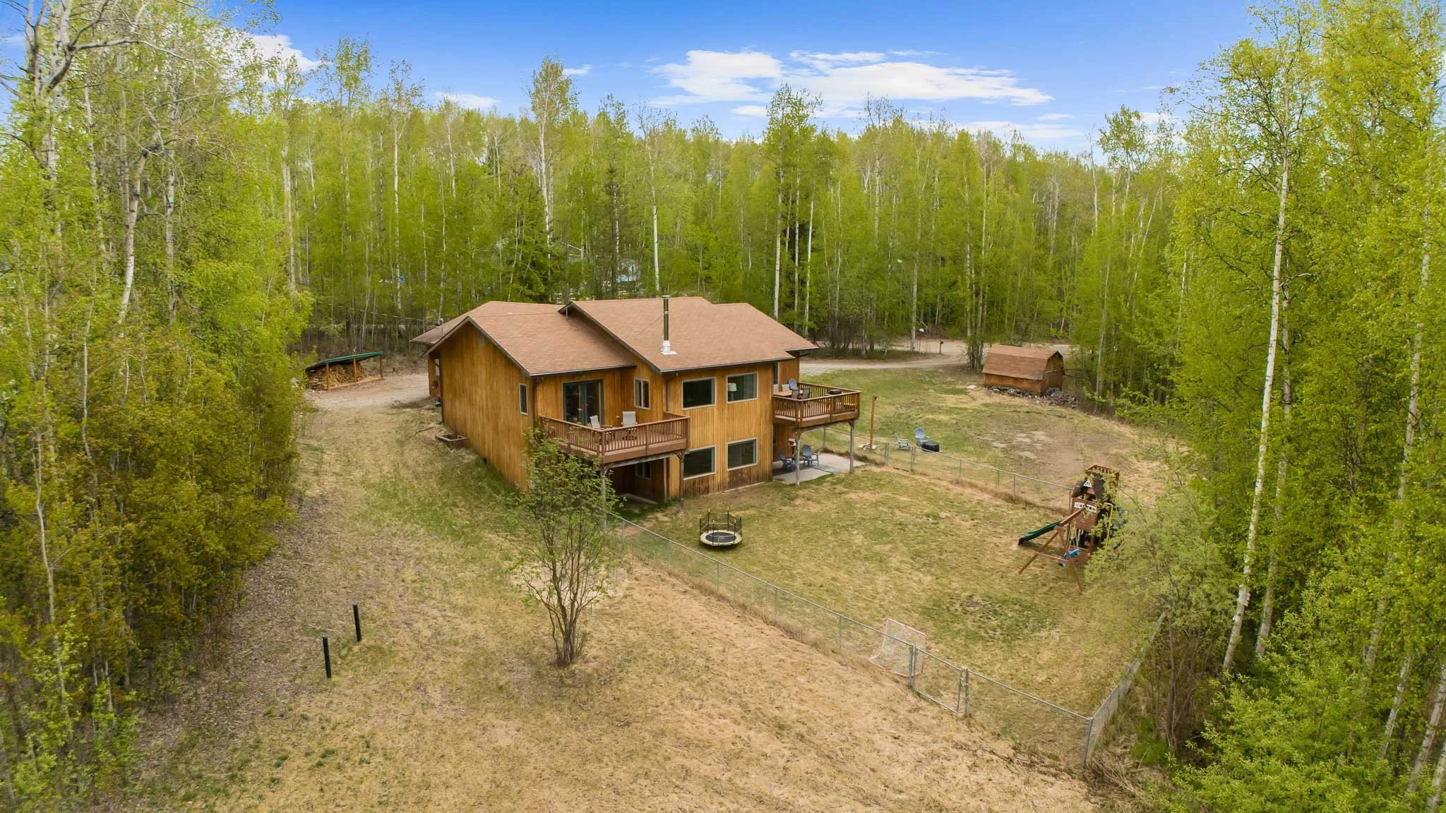 A two-story wooden house with a brown roof and large deck in a wooded area with green trees and a grassy yard.