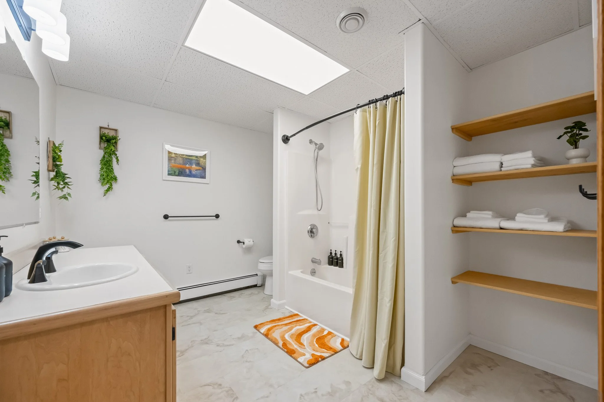 A clean, bright bathroom with a vanity sink and faucet in the front left, a bathtub with a showerhead and yellow shower curtain in the center, a wooden shelf with rolled towels and a potted plant on the right, white walls with hanging green plants an
