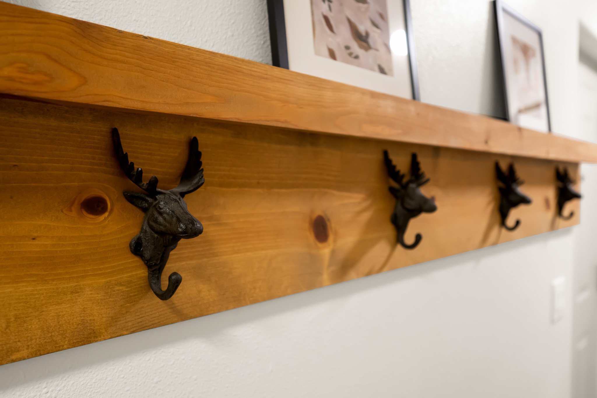 Decorative wall shelf made of wooden plank with black metal deer head hooks, tied to a wall with framed artwork in the background.