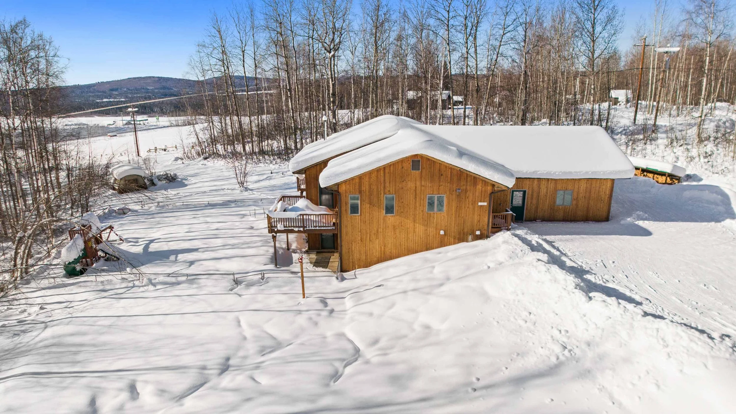 A wooden house with a snow-covered roof, surrounded by snow, trees, and distant hills on a clear winter day.