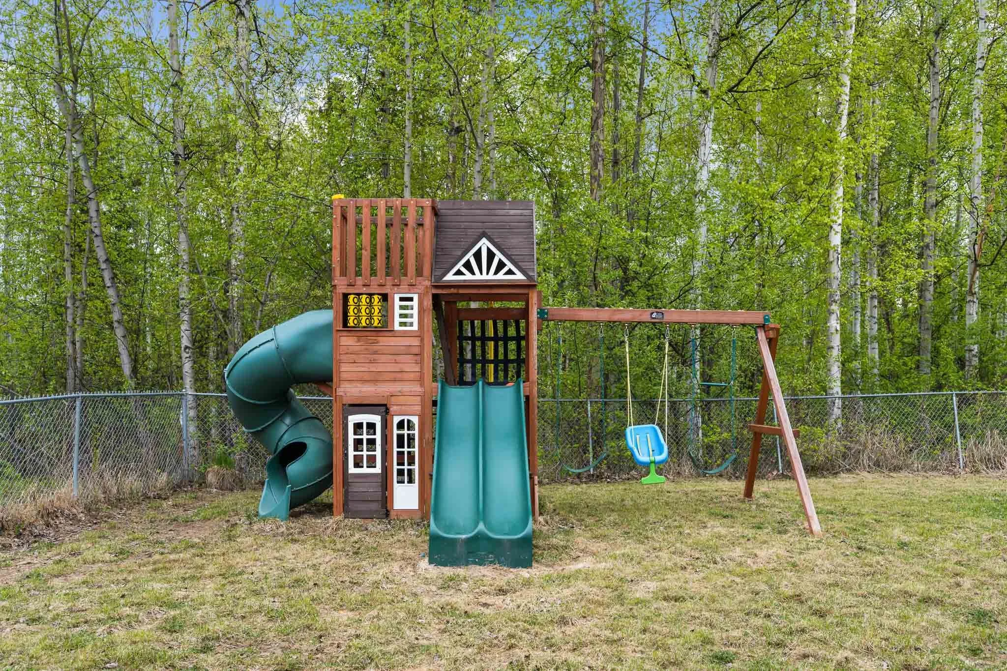 A backyard playground with a wooden playhouse, a green slide, a blue swing set, and a green plastic baby swing, surrounded by a chain-link fence and tall trees with green leaves.