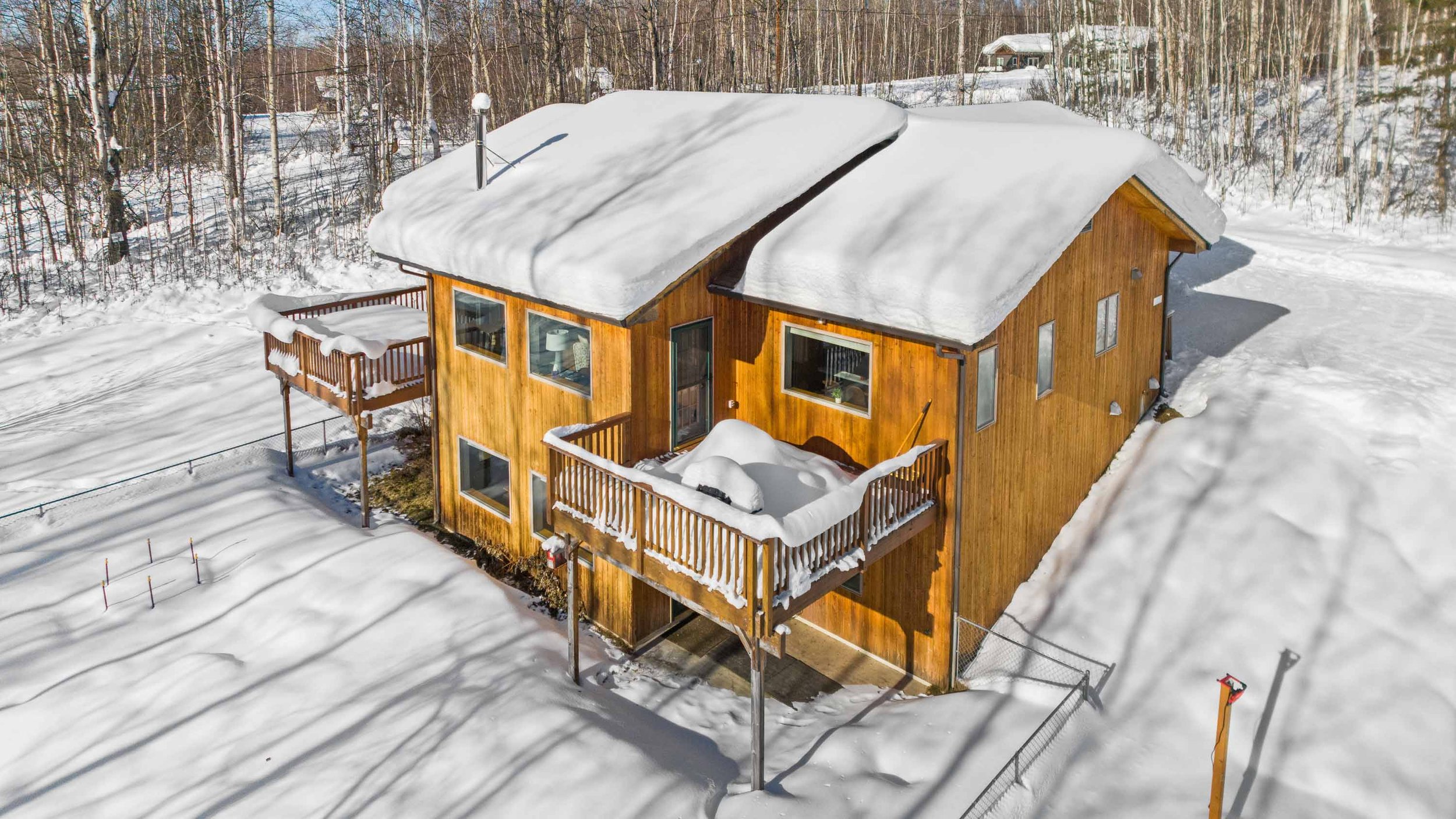 A wooden house with snow-covered roof and balconies, surrounded by snow and trees during winter.