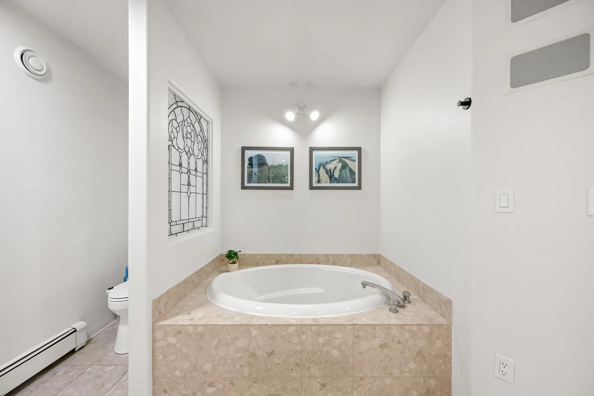 Bathroom with a built-in bathtub, artwork on the walls, a small potted plant, and a window with stained glass.