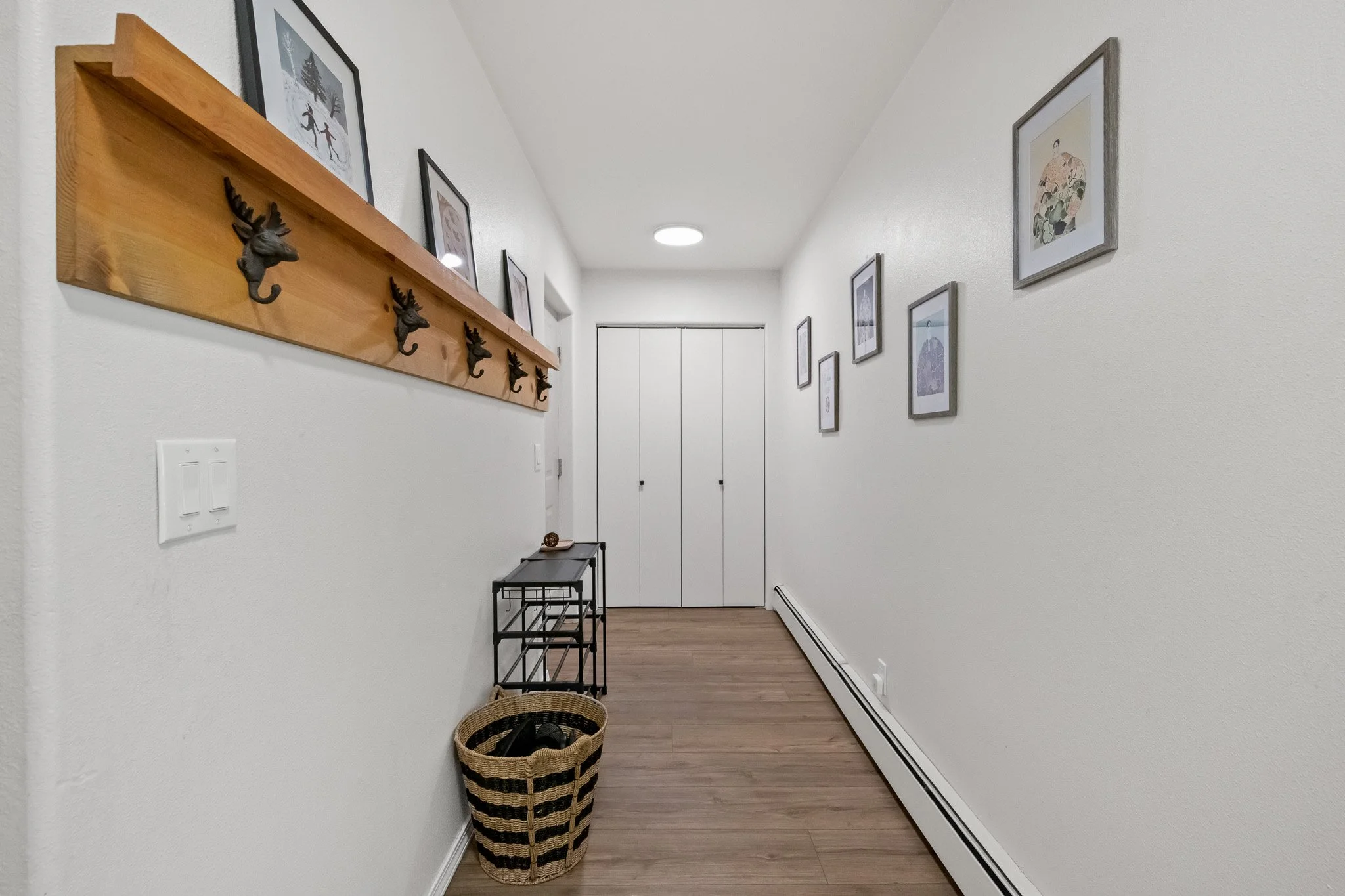 Hallway with framed pictures on the wall, a wooden coat rack with hooks, a small black table, a woven basket, and baseboard heating.