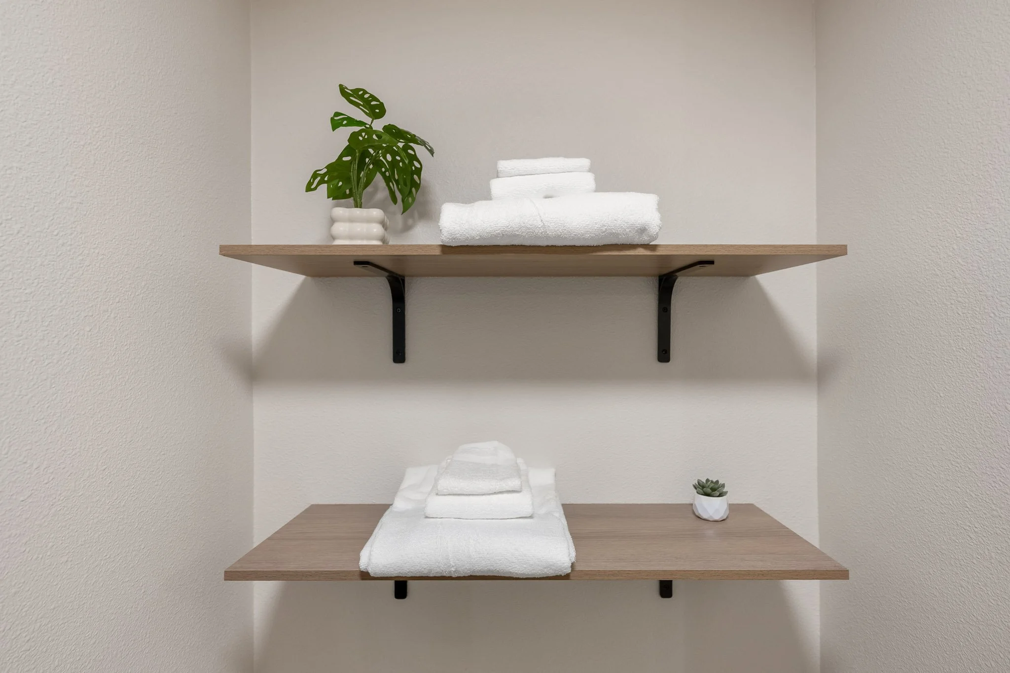 A minimalist bathroom shelf setup with a potted monstera plant and folded white towels on two wooden shelves against a white textured wall.