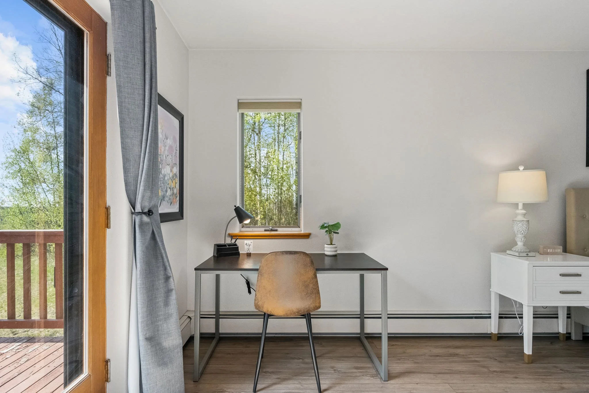 Interior view of a room with a sliding glass door, a desk with a lamp, a chair, and a window with trees outside.