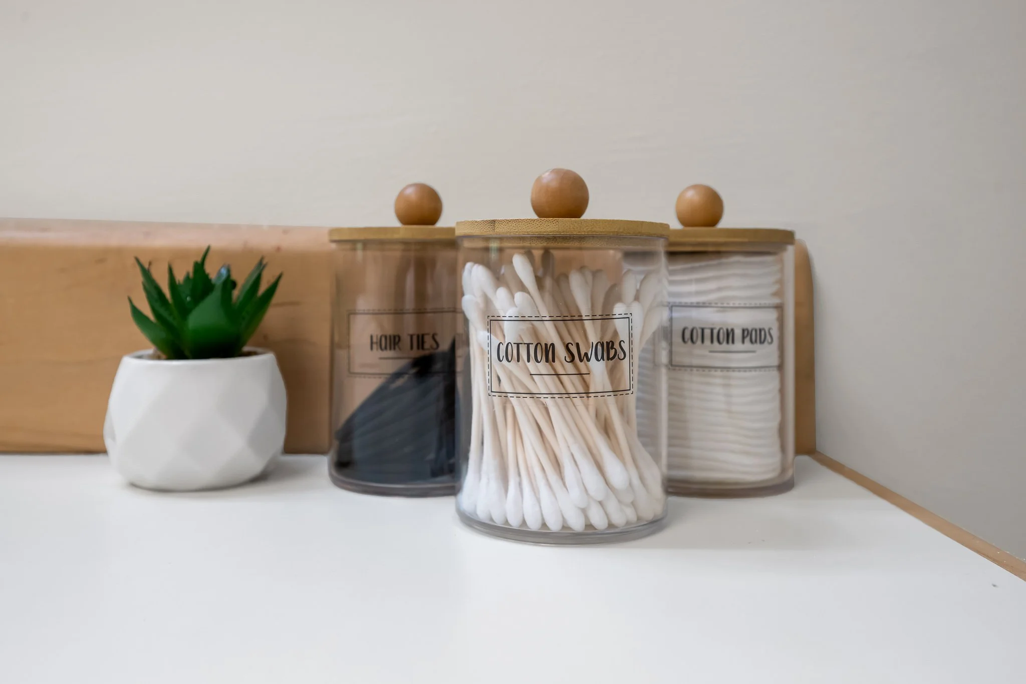 A wooden cabinet with three labeled containers holding hair ties, cotton swabs, and cotton pads. To the left, a white geometric pot contains a green succulent plant.