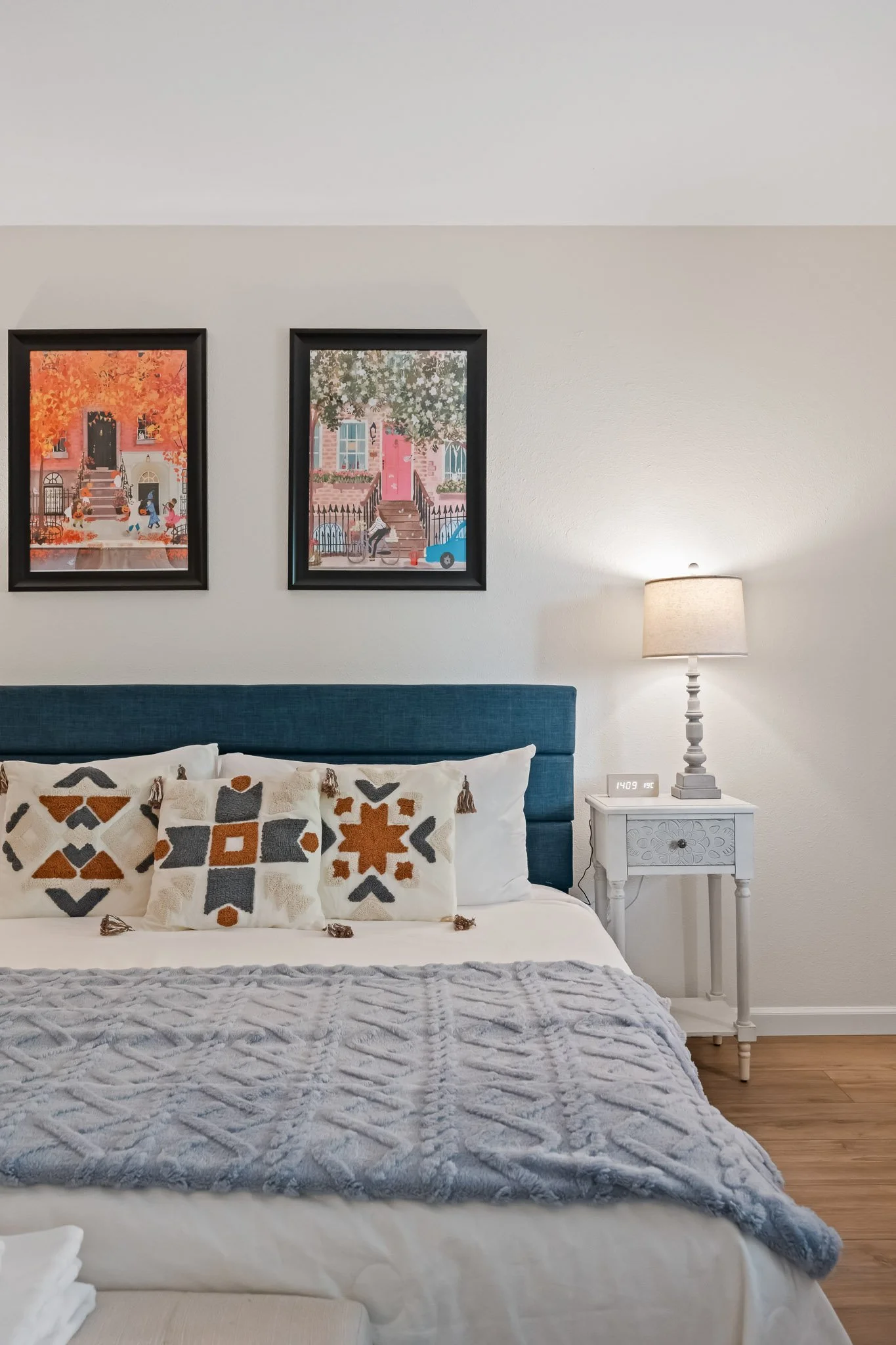 A cozy bedroom with a white wall and two framed colorful illustrations of houses and trees hanging above a bed with white and patterned pillows. There is a nightstand with a lamp and a digital clock showing 14:09.