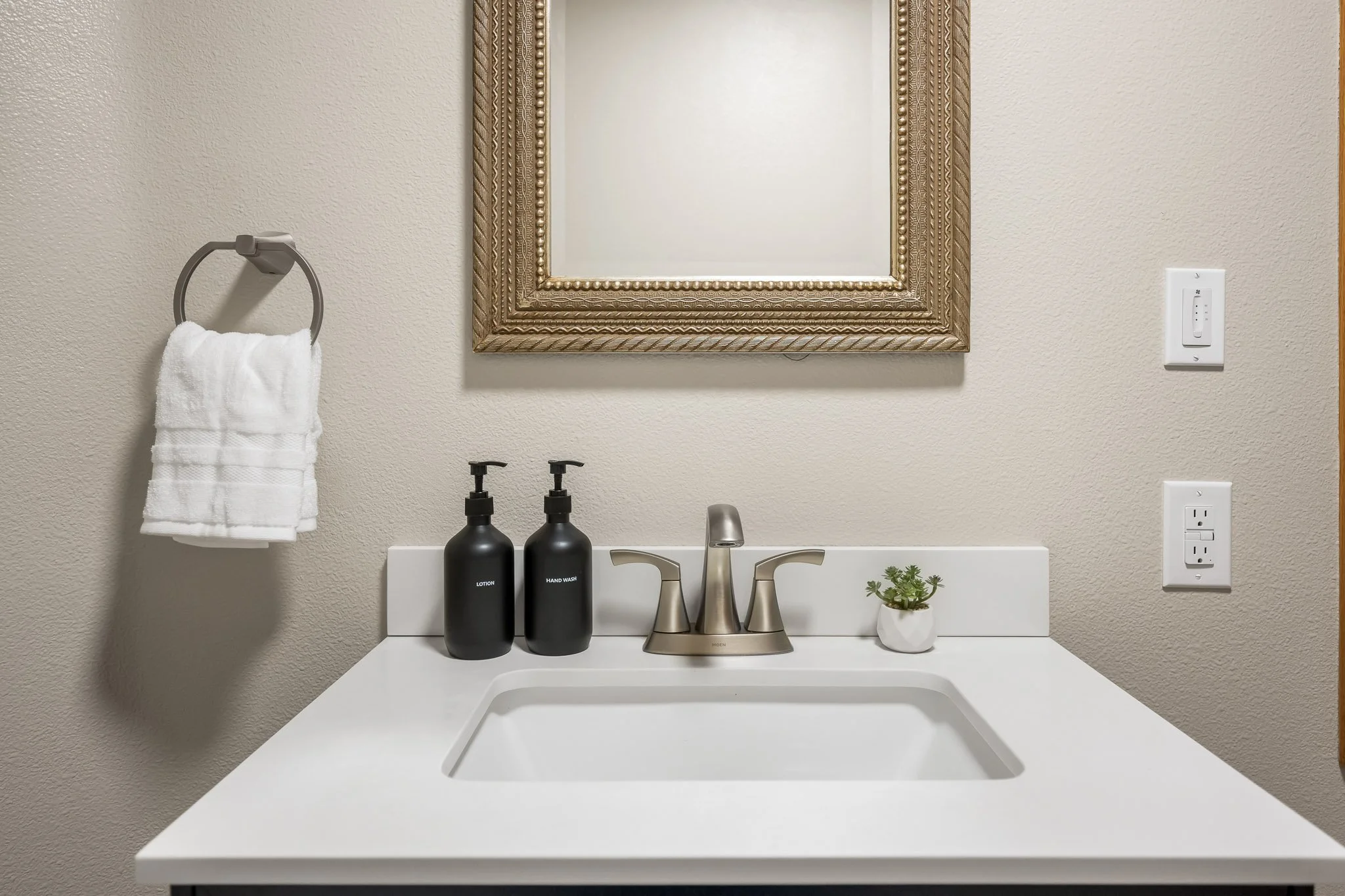 Bathroom vanity with a large framed mirror, two black bottles labeled 'Lotion' and 'Hand Wash,' a small potted plant, a double-handle faucet, a white countertop, a towel hanging on a silver ring, and electrical outlets.