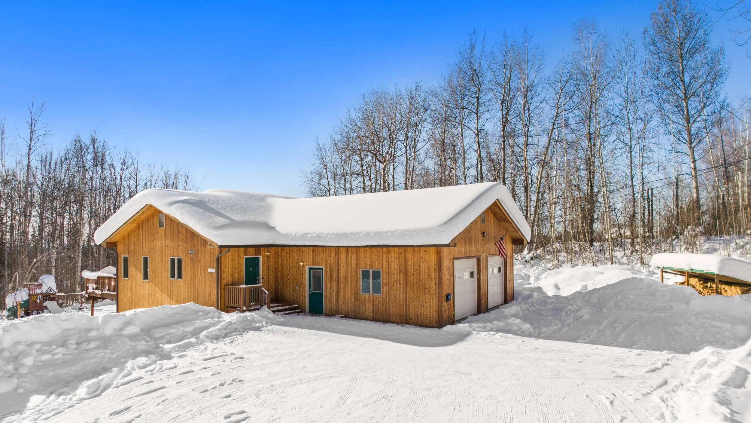 A wooden house with a snow-covered roof, surrounded by snow, and a forest of bare trees in the background on a bright, clear day.