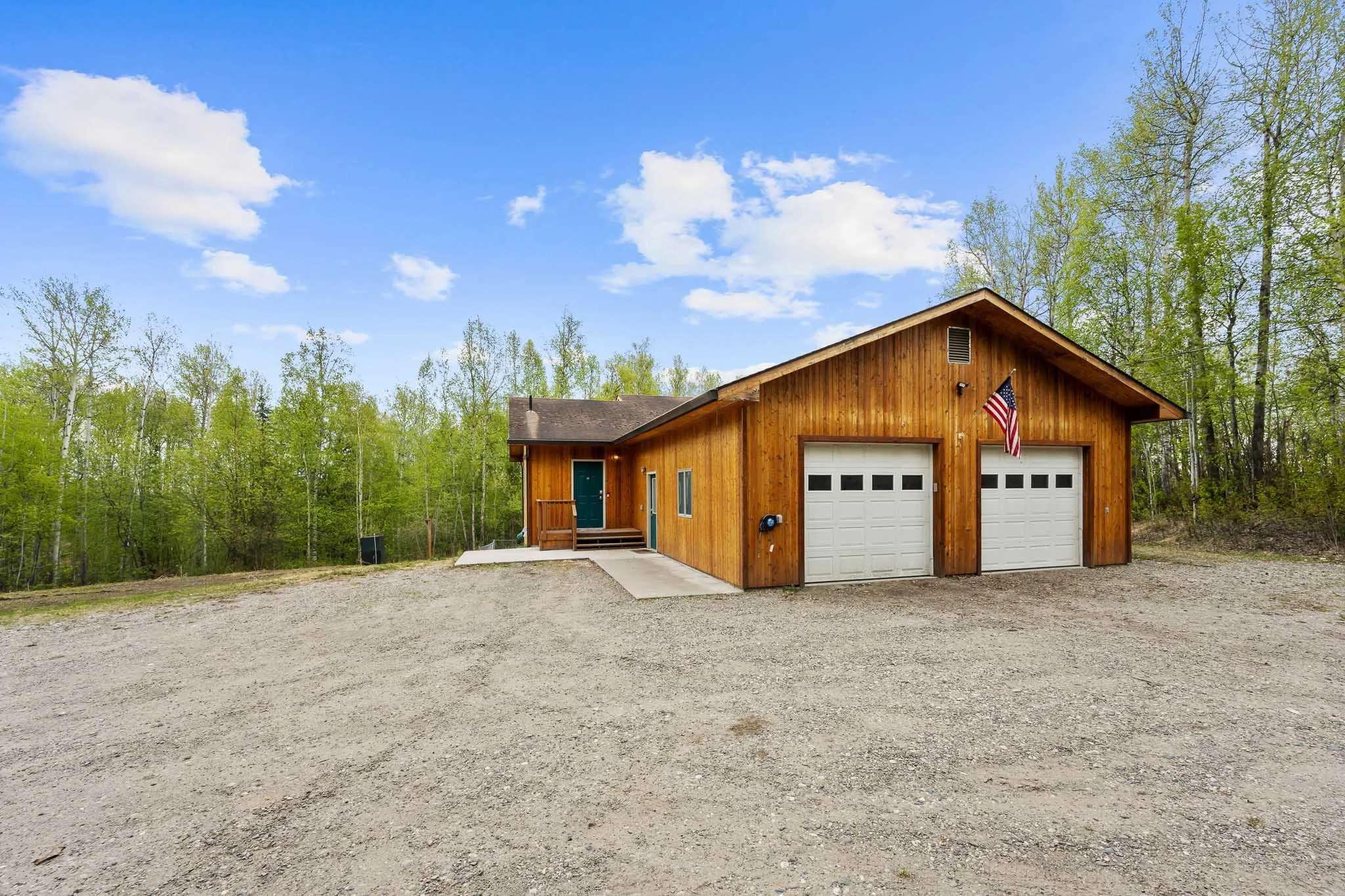 A wooden garage with two white doors and an American flag hanging on its side, surrounded by green trees and a gravel driveway, under a partly cloudy blue sky.