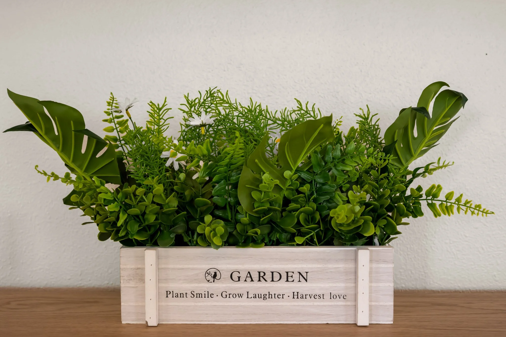 Arrangement of green artificial plants in a white wooden box with the words 'GARDEN' and a quote about planting and loving on it, placed on a wooden surface against a plain wall.
