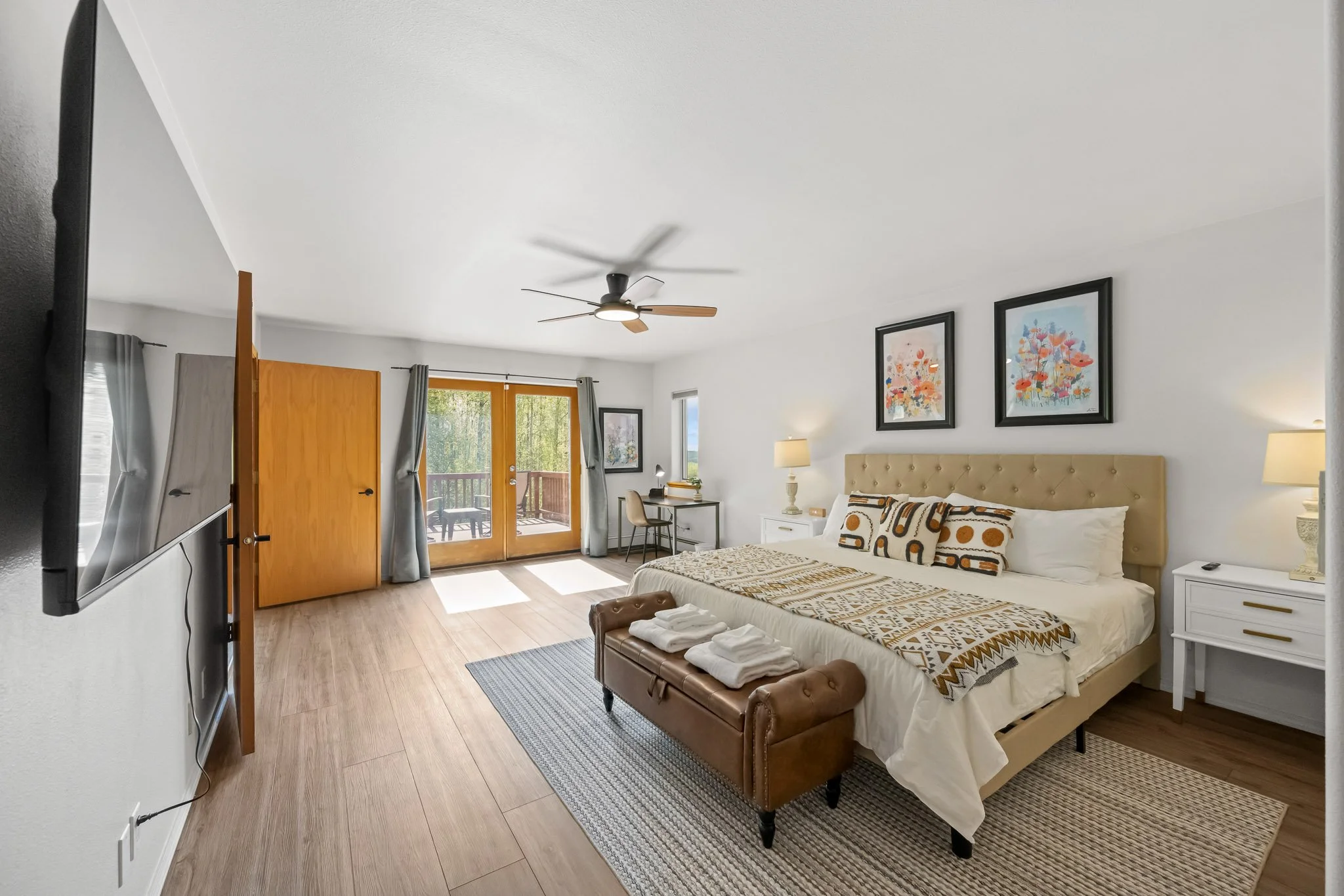 Interior of a bedroom with a large bed, framed colorful artwork on the wall, nightstands, lamps, a small desk with a chair, and a balcony door with curtains.