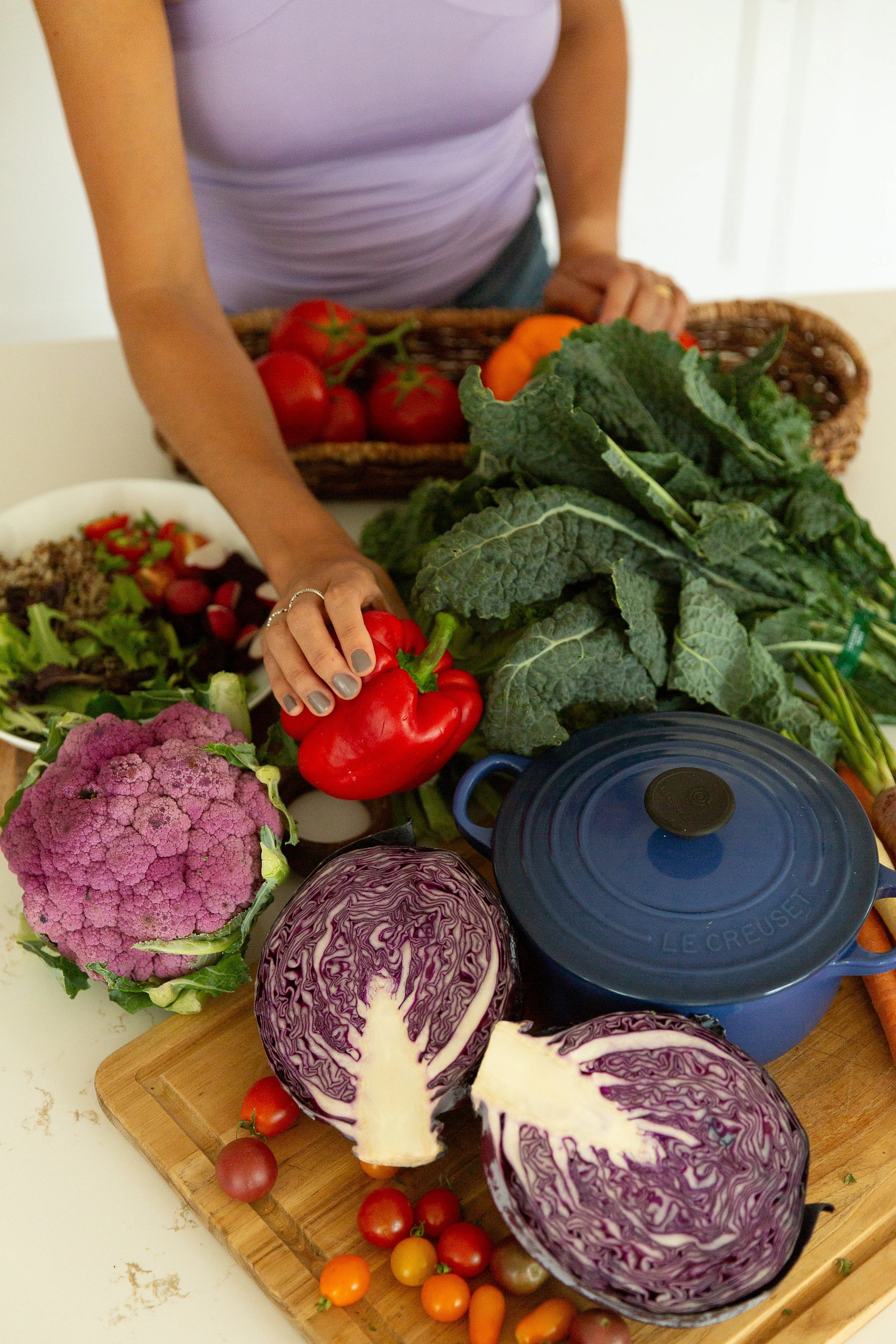 A person selecting a red bell pepper from a display of fresh vegetables, including cherry tomatoes, cauliflower, purple cabbage, kale, and tomatoes, on a wooden board and in a basket.