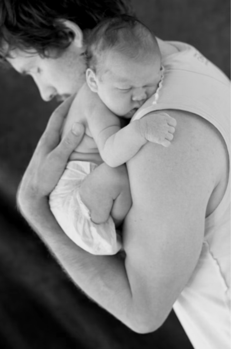 Black and white photo of a parent holding a sleeping newborn baby close, with the child's face resting on the parent's shoulder.