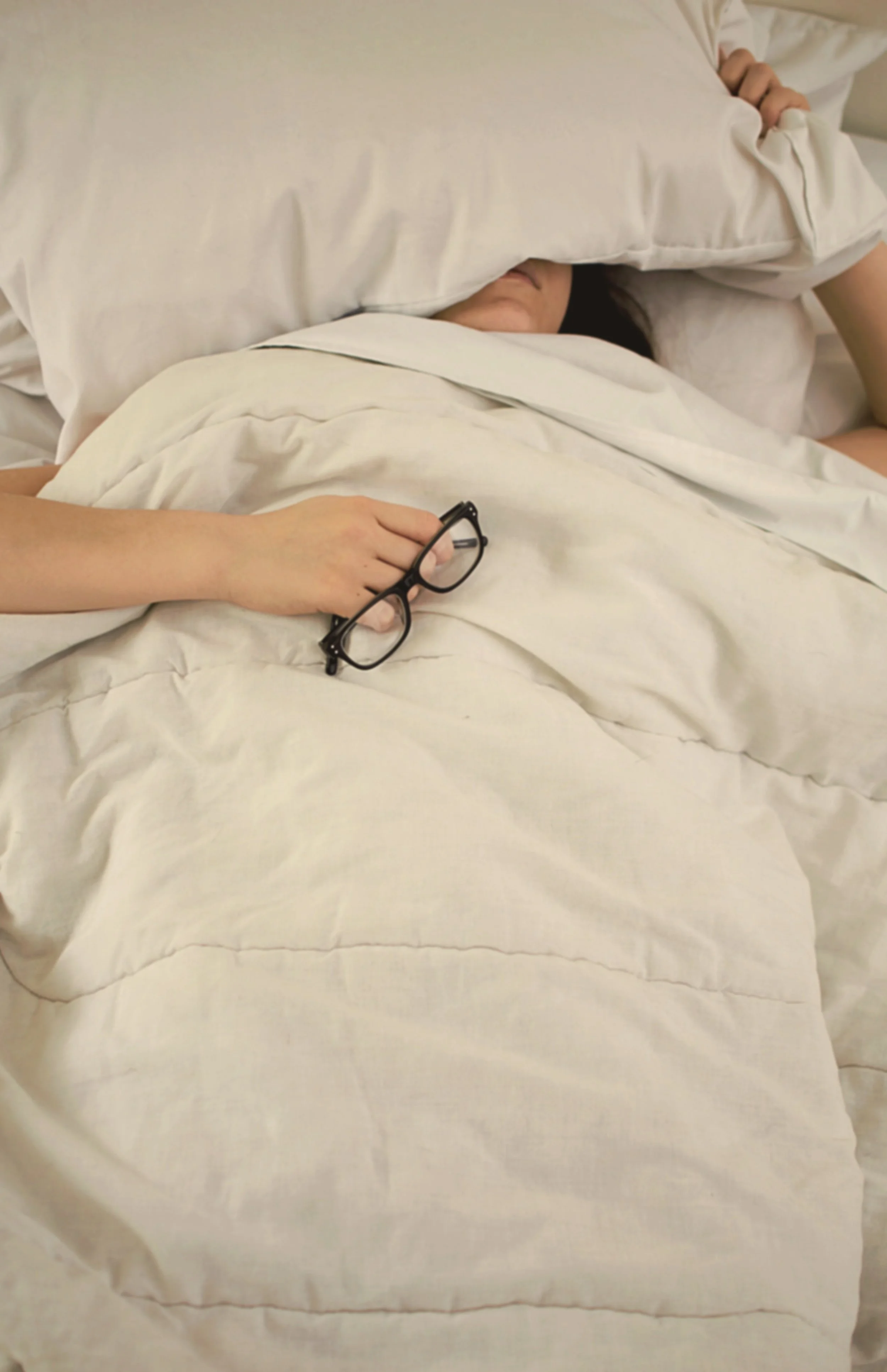A person lying in bed, partially covered by white sheets and pillows, holding a pair of black glasses in their left hand, with their face partially visible between the pillow and sheets.
