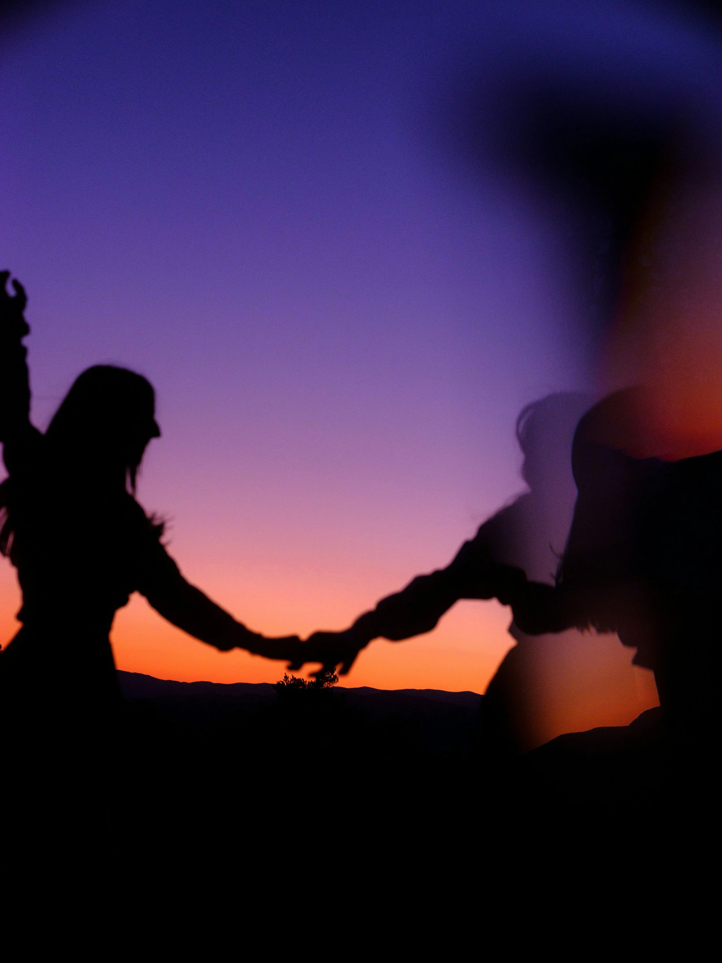 Silhouettes of two people holding hands during a sunset with a colorful sky in the background.