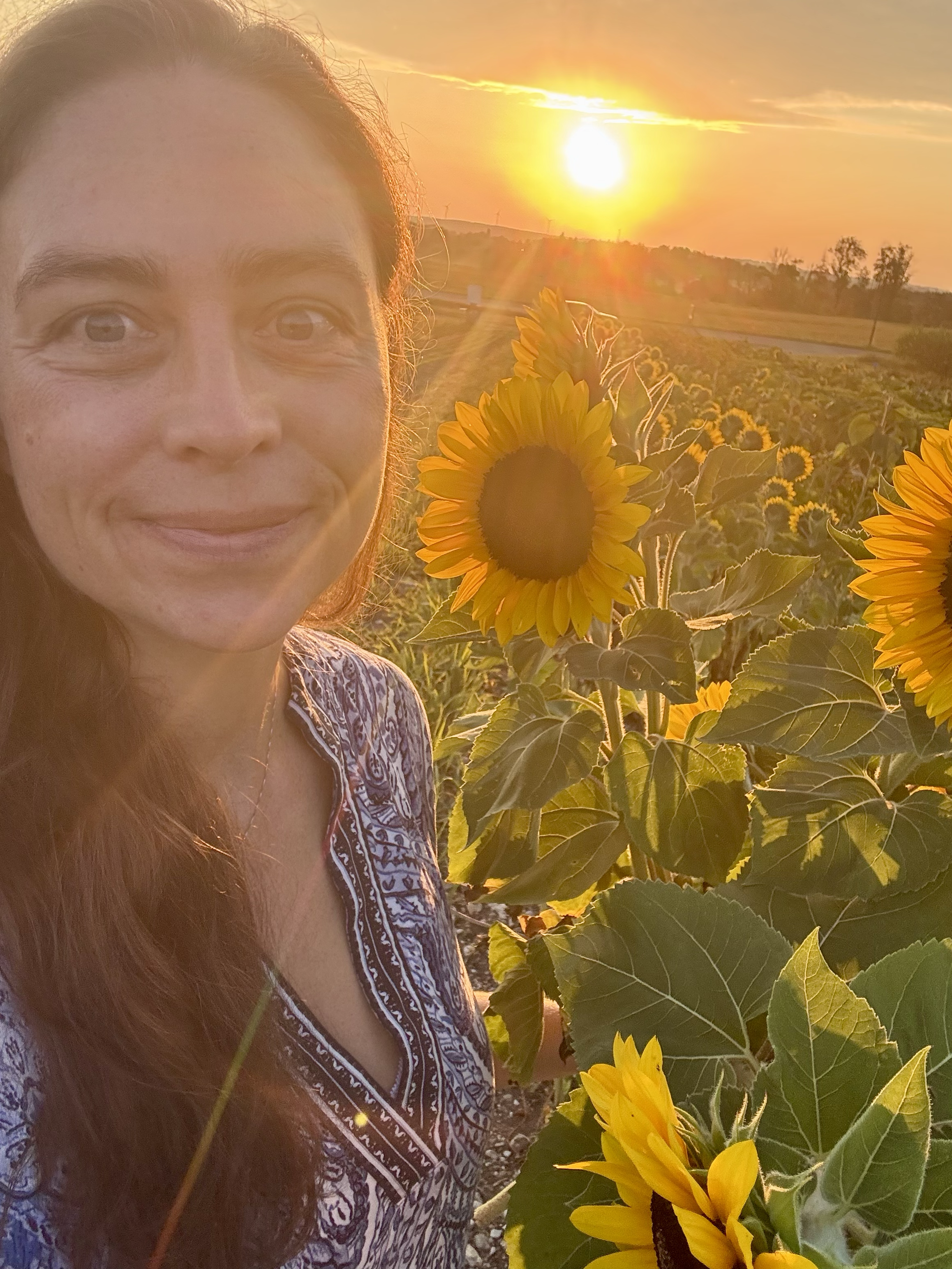 A woman with long brown hair smiling in a sunflower field at sunrise.