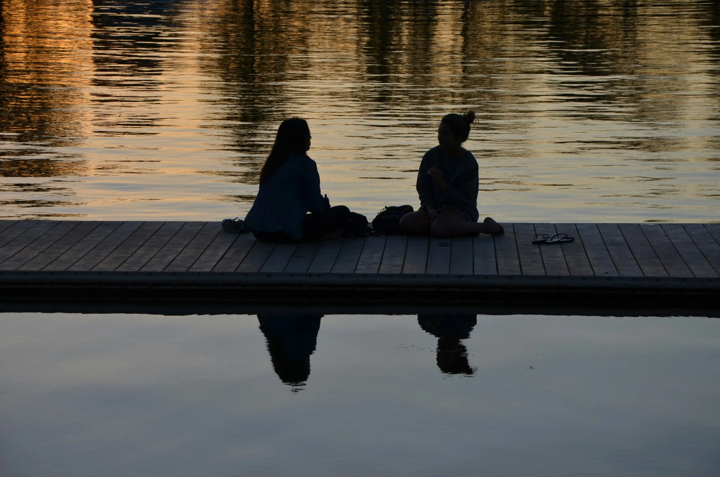 Two silhouetted women sitting on a dock by a calm lake during sunset, with their reflection visible in the water.