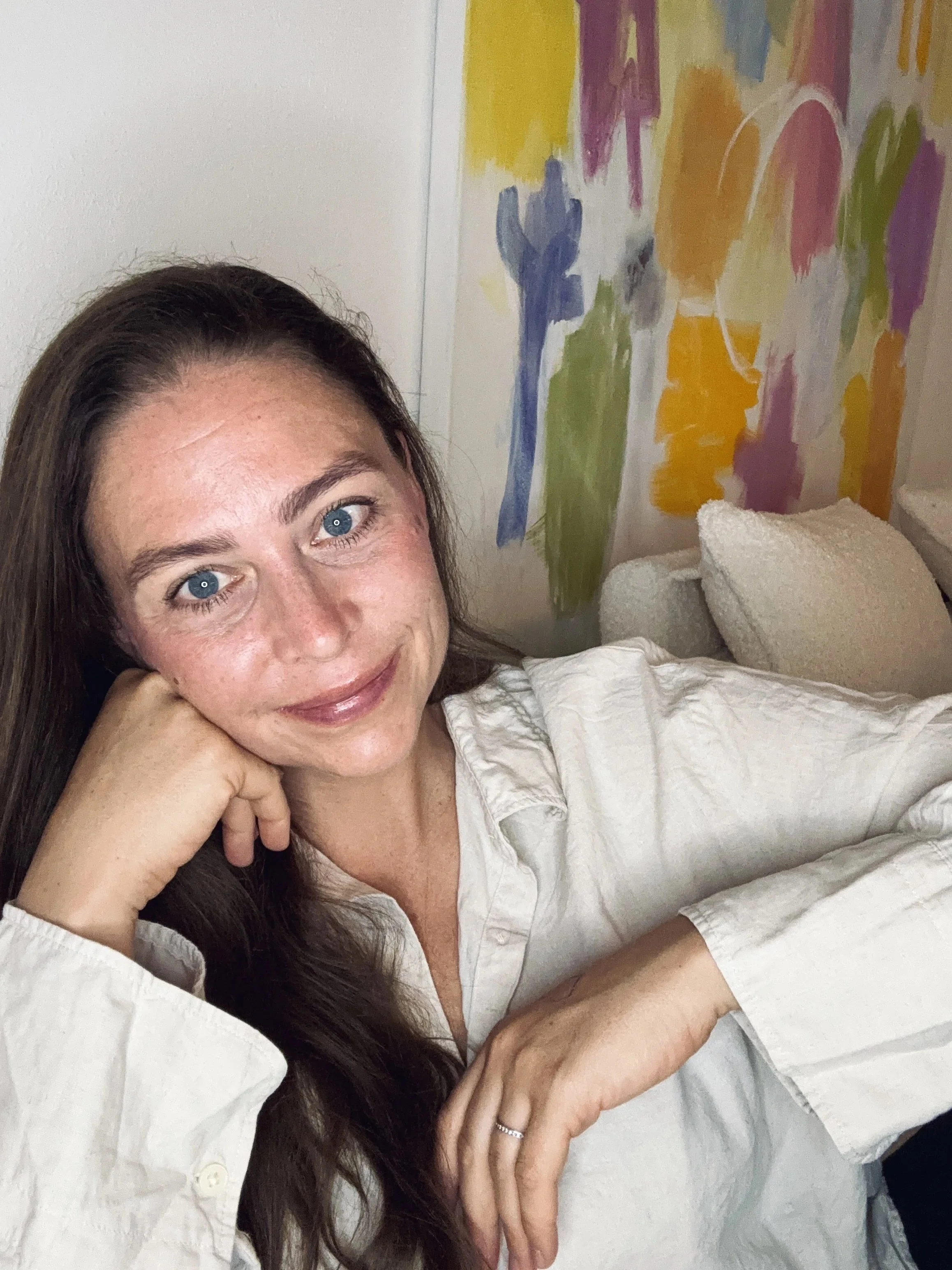 A woman resting her head on her right hand, smiling at the camera, with long brown hair, wearing a white blouse, next to a bed with white sheets and a colorful abstract painting in the background.