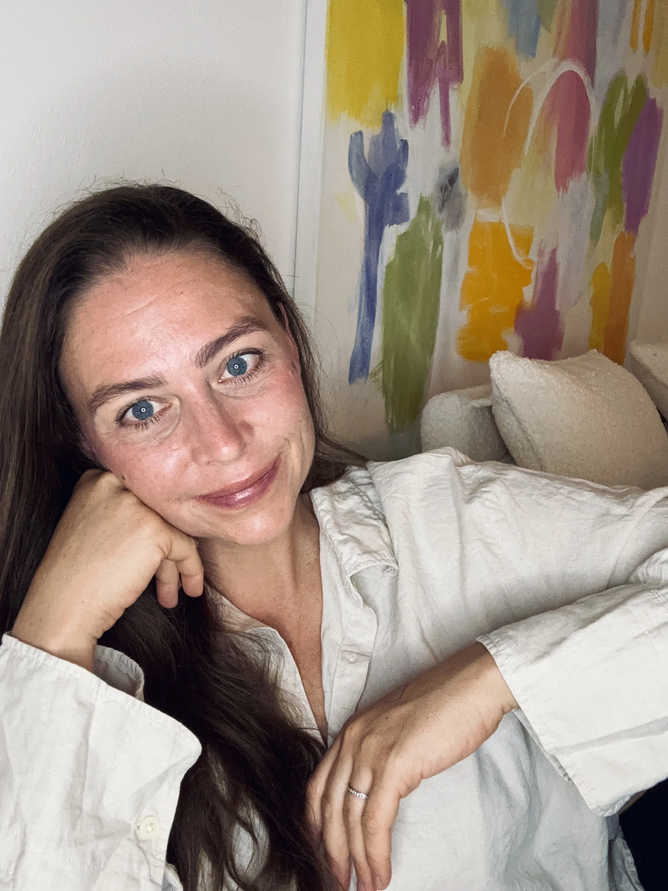 A woman with long brown hair, blue eyes, and fair skin, smiling, with her head resting on her hand. She is wearing a white shirt. In the background, there is an abstract colorful painting with pink, yellow, blue, green, orange, and purple colors, and a folded white towel or blanket on the bed.