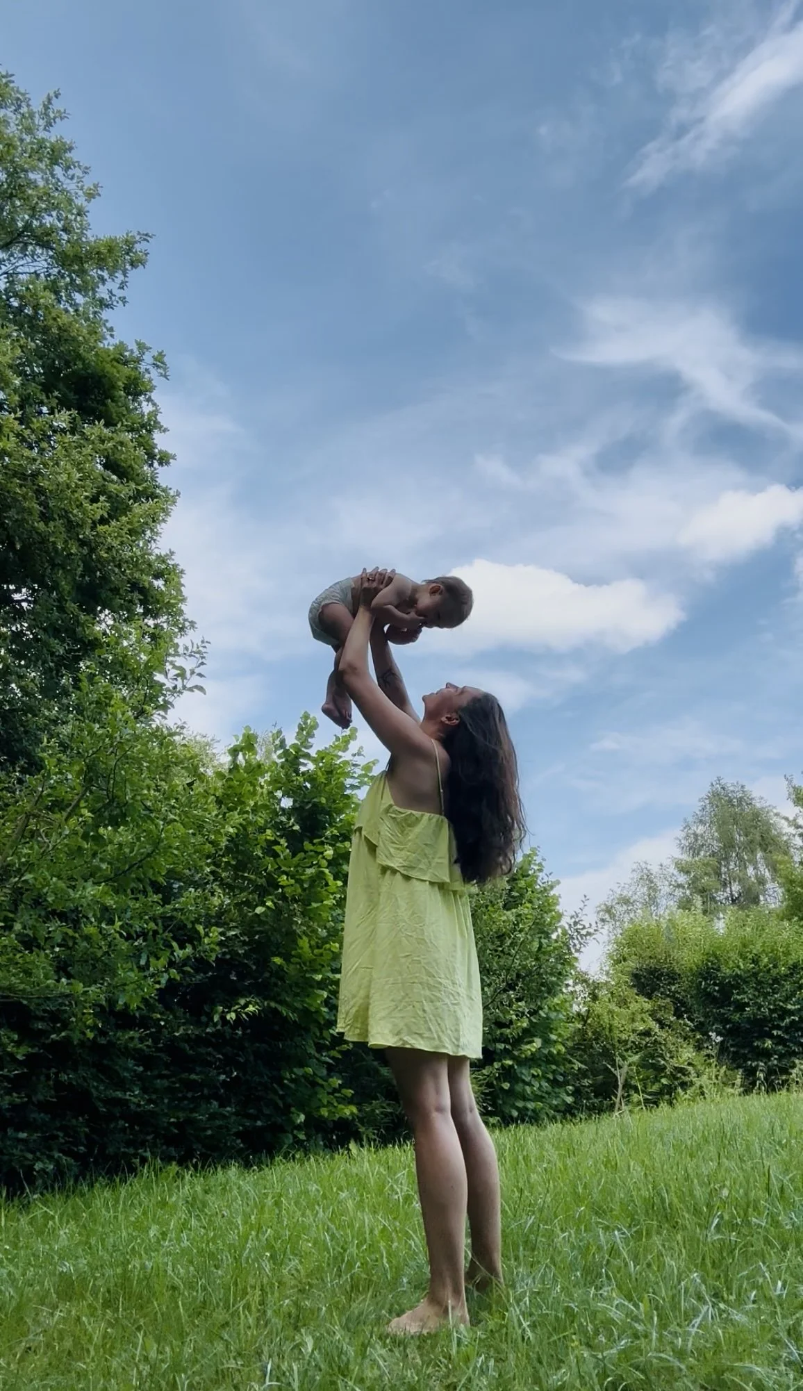 A woman in a greenish dress is holding a young child up in the air outdoors in a green field with trees and a blue sky with clouds overhead.