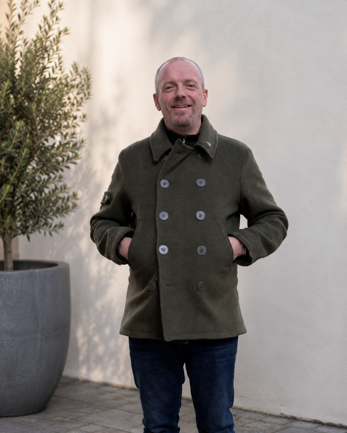 Smiling man with a shaved head and goatee wearing a dark green double-breasted coat and blue jeans, standing outdoors near a large potted plant against a plain light-colored wall.