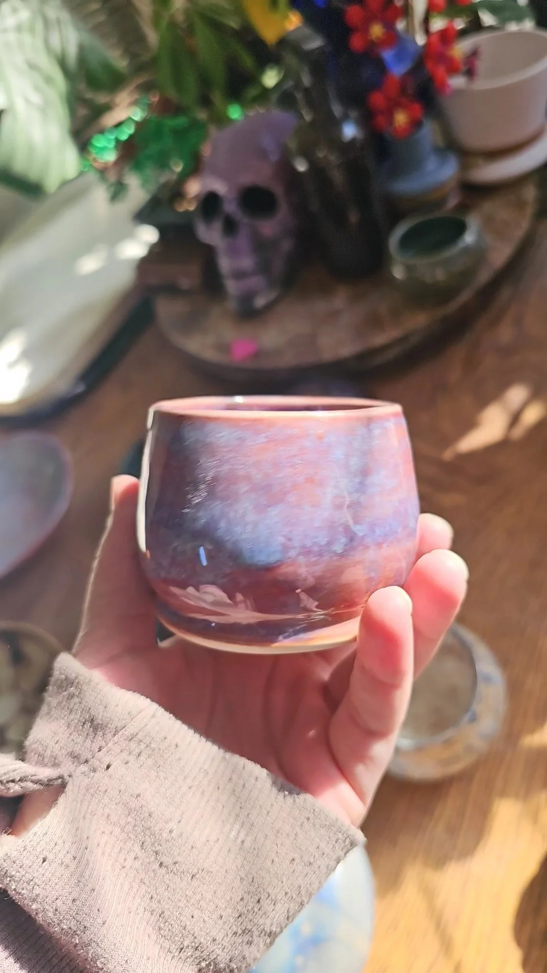 A hand holding a pink and purple ceramic cup in a room with various pottery items and a black skull decoration on a table.