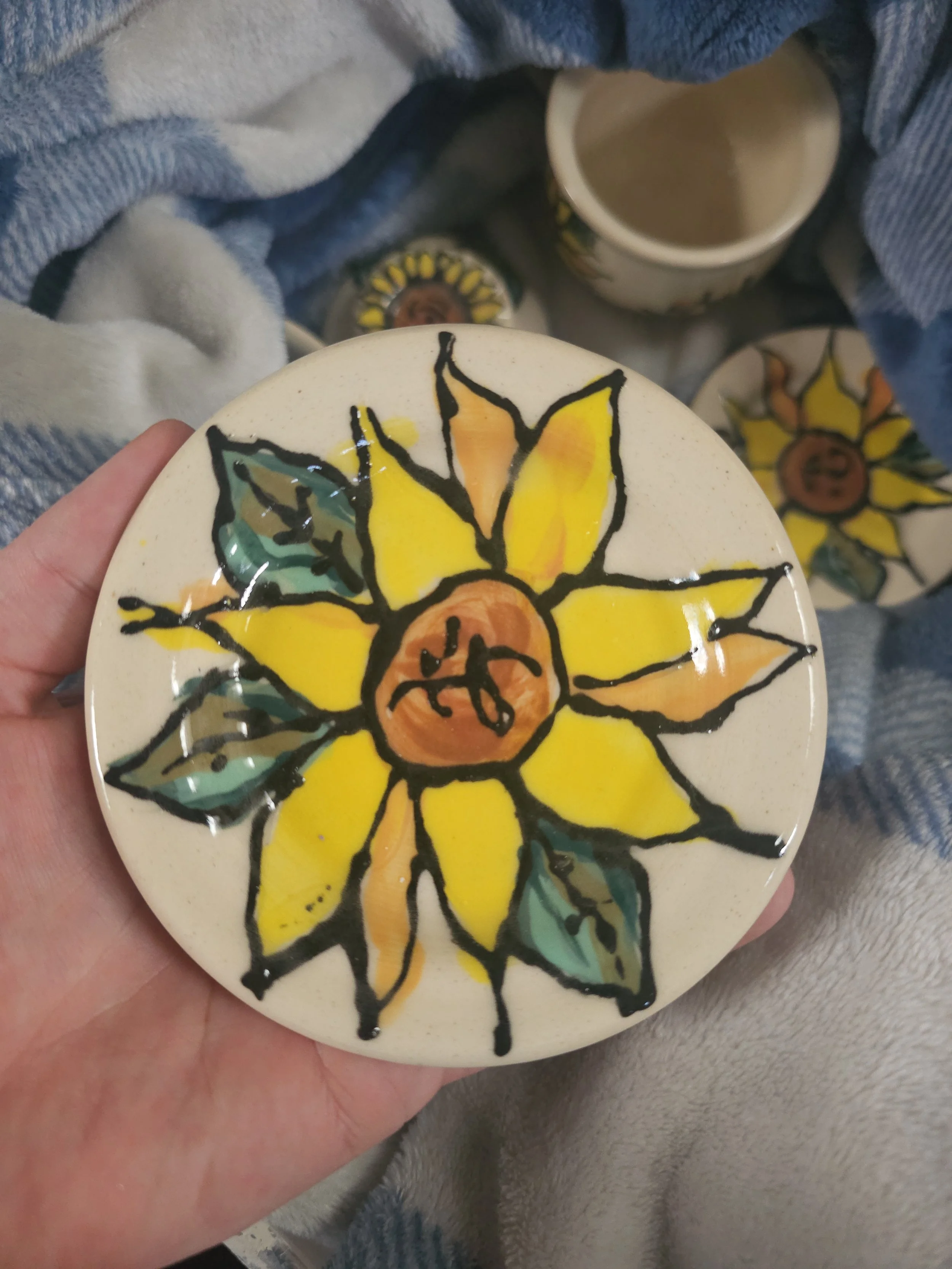 Hand holding a ceramic plate with a hand-painted yellow and orange flower with green leaves.