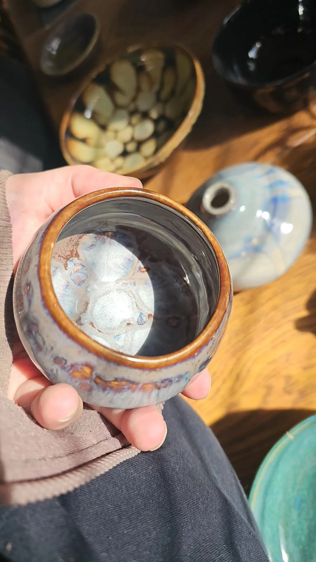 A person holding a ceramic cup with an iridescent glaze, reflections visible inside the cup. In the background, there are other ceramic vessels, including a mottled brown and beige bowl and a tall, light blue and white vase with a narrow opening. The