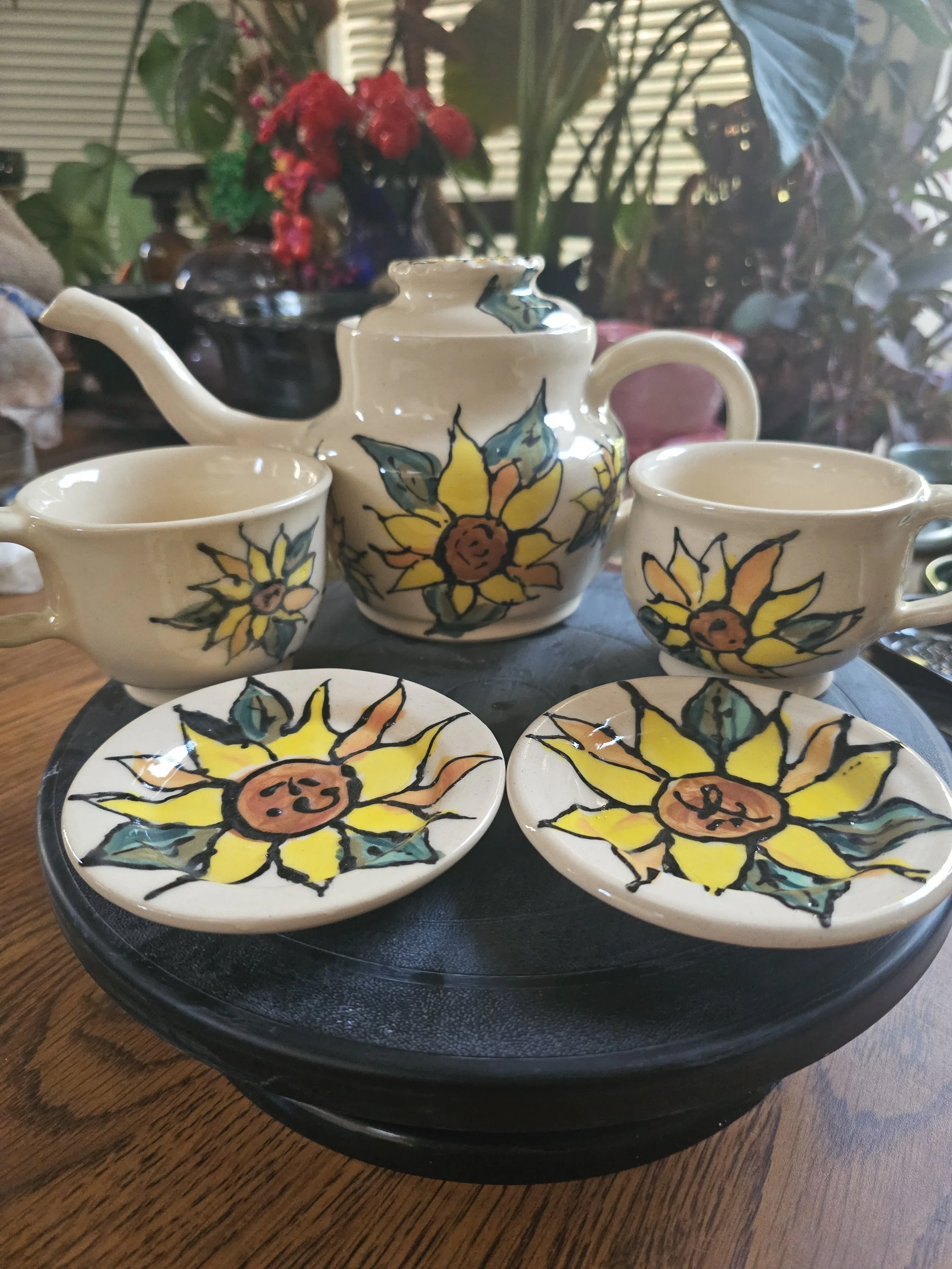 Ceramic teapot, two matching cups, and two small plates with sunflower designs, arranged on a black tray on a wooden table. In the background, there are various potted plants and flowers.