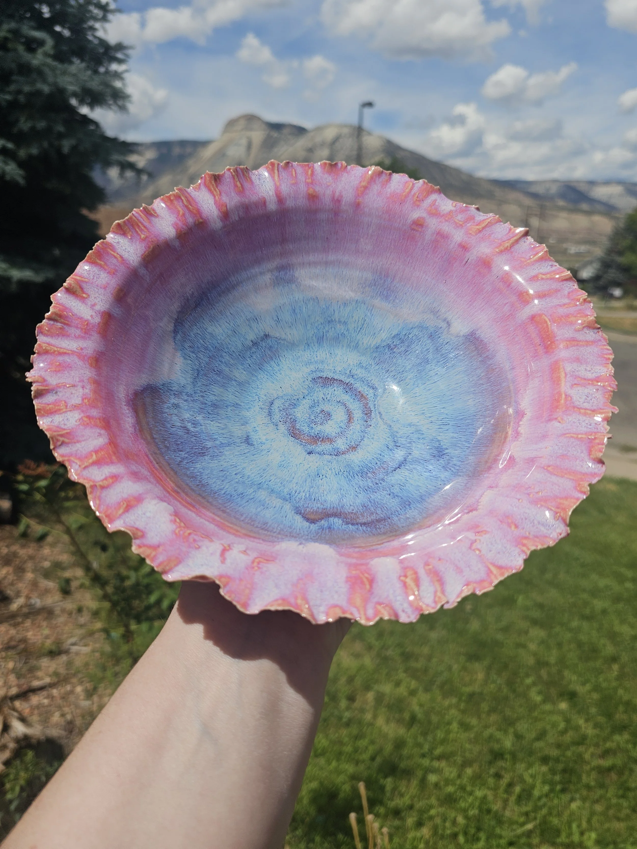 A colorful ceramic bowl with a blue and pink pattern, outdoors with a mountain landscape and a partly cloudy sky in the background.