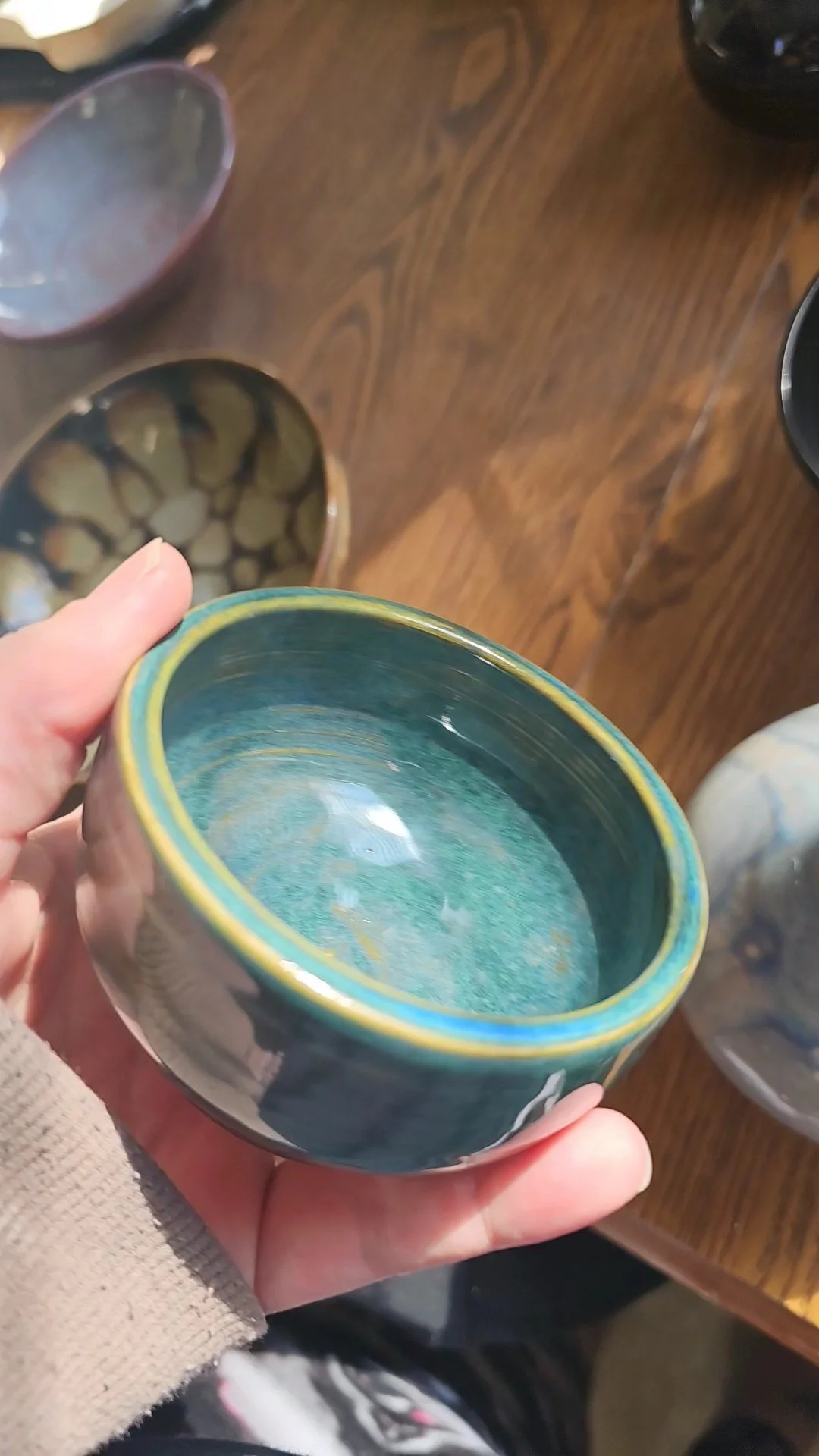 A person holding a small ceramic bowl with a blue interior and a yellow rim, surrounded by other bowls on a wooden surface.