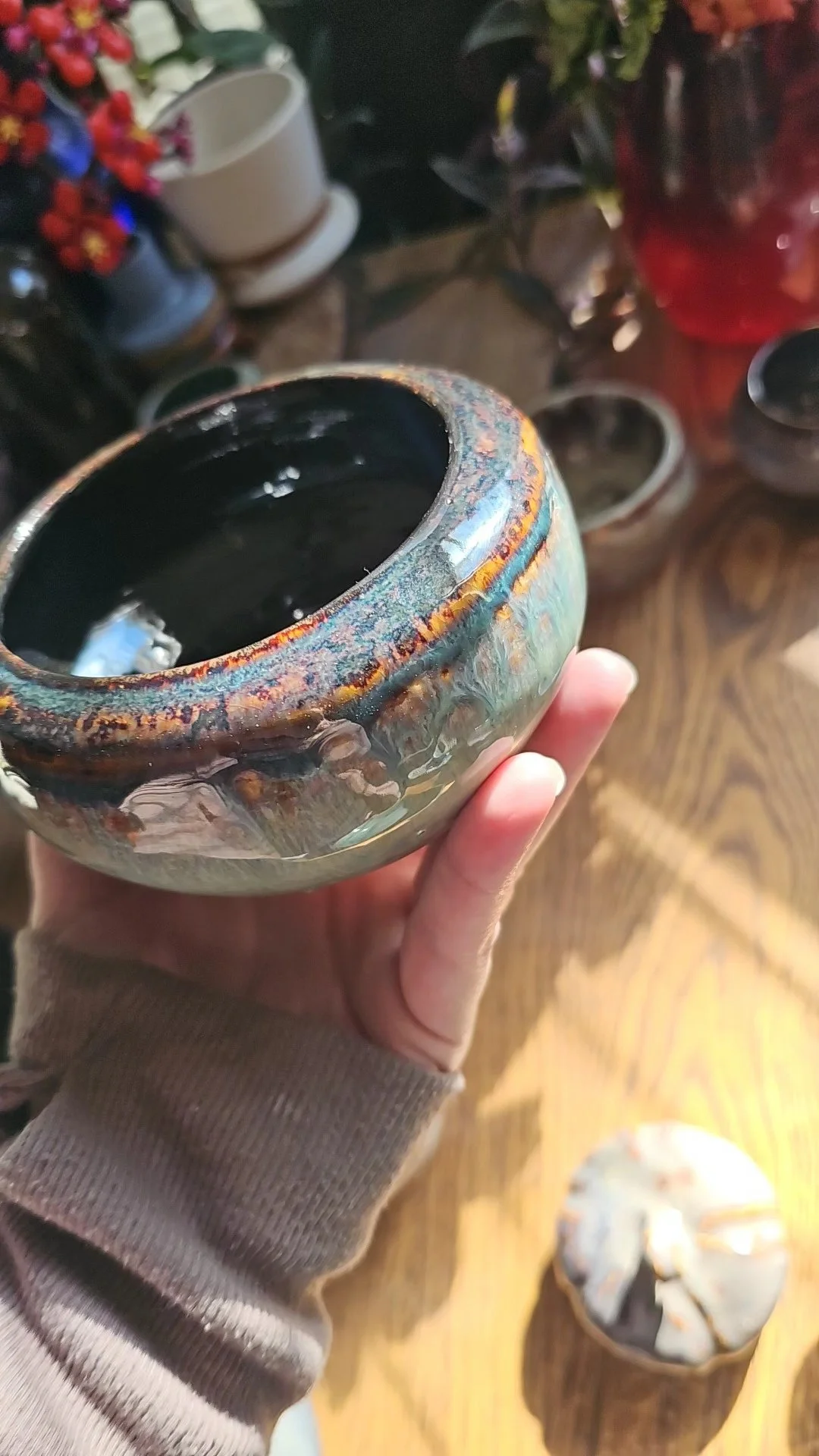 A person holding a small, round, glazed ceramic bowl with a dark interior, situated in a store surrounded by various other pottery and plant pots.