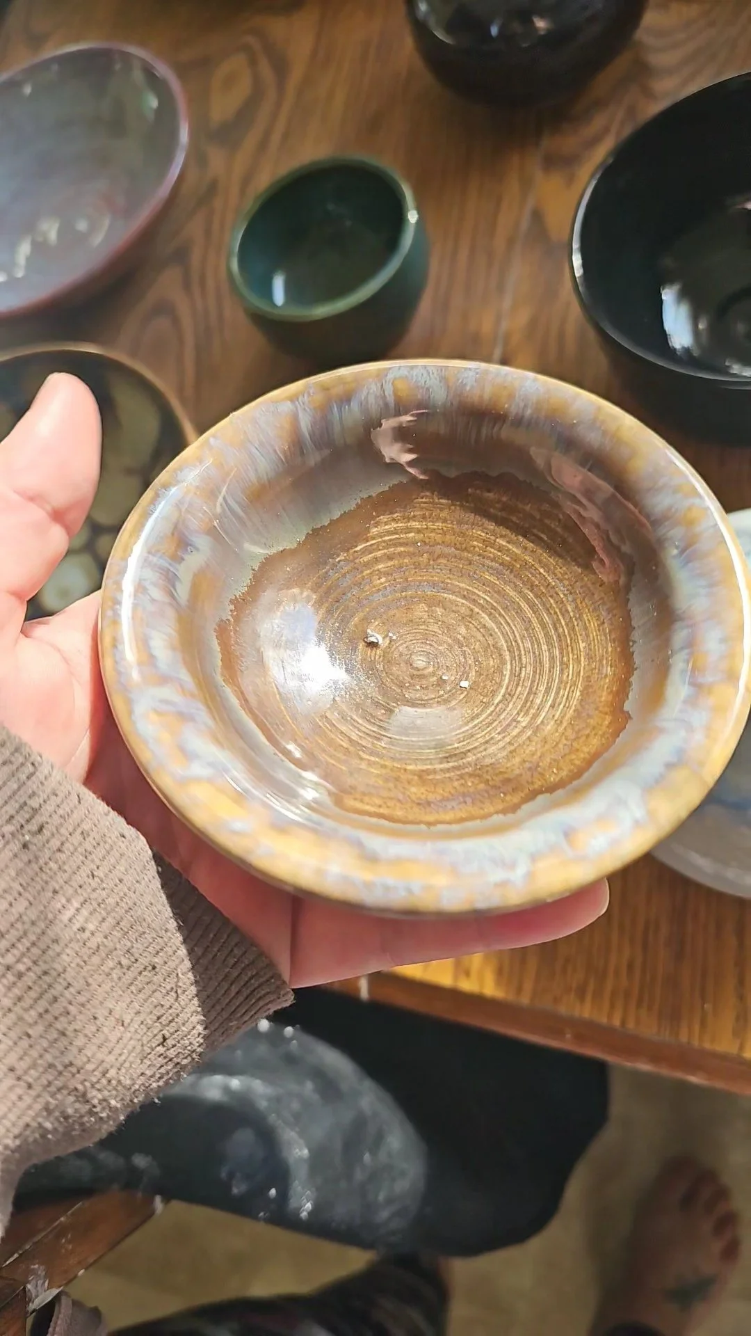 A hand holding a ceramic bowl with a glossy, marbled pattern in earthy tones, on a wooden table with various other ceramic bowls around.