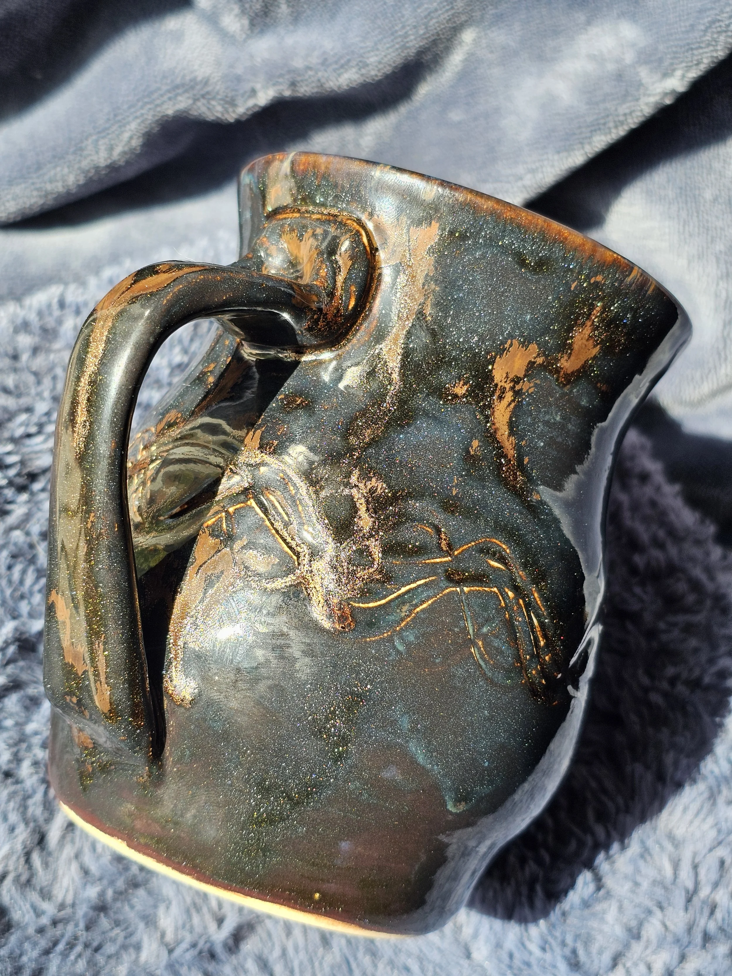 Close-up of a decorative glass mug with swirling black, copper, and iridescent patterns, held against a grey fabric surface.