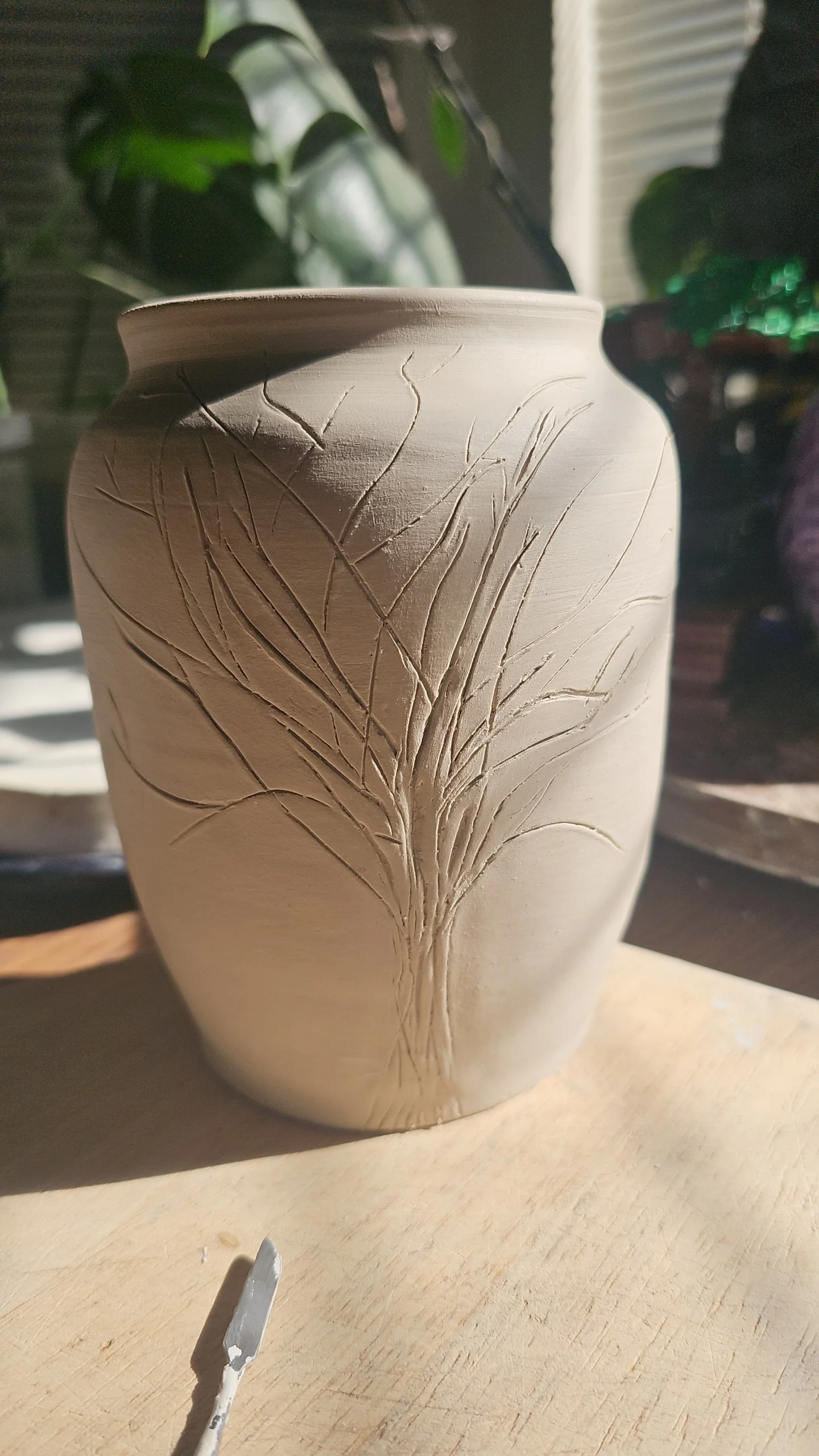 A ceramic vase with engraved leaf patterns, placed on a wooden surface with a plant in the background.