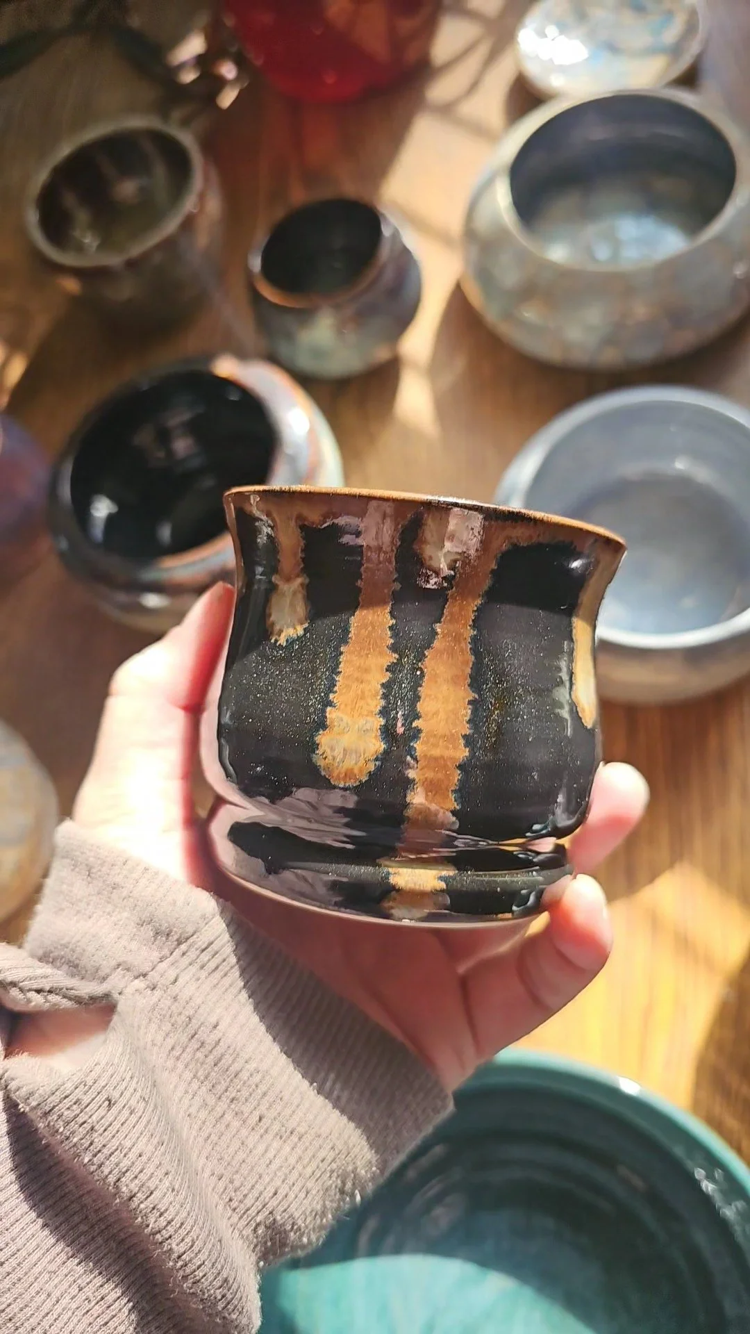 A person holding a small black-glazed ceramic bowl with brownish streaks, surrounded by other similar ceramic bowls on a wooden surface.