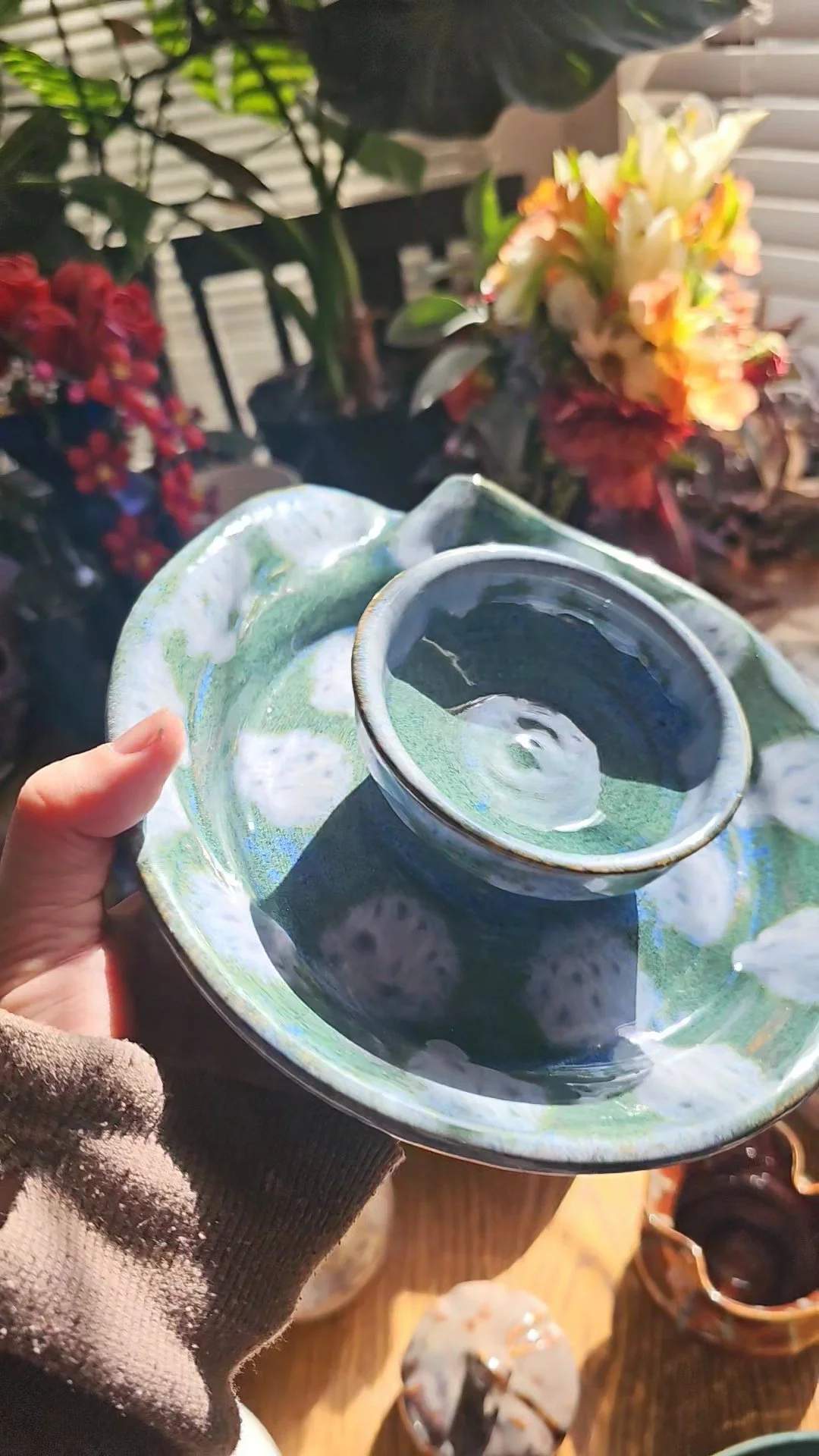 A person holding a ceramic dish with a smaller bowl on top, filled with water, against a background of colorful flowers and plants.