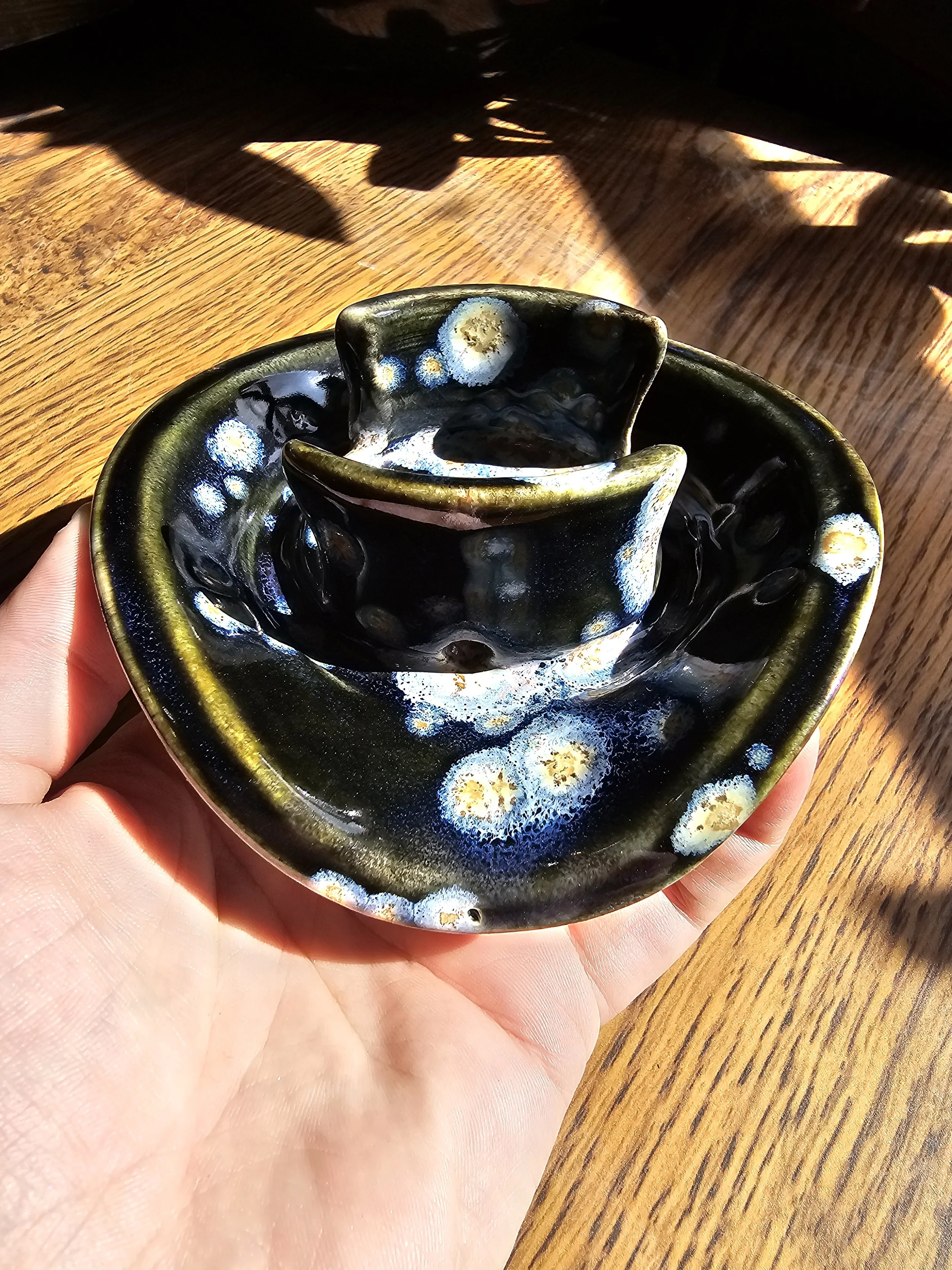 A hand holding a small black ceramic bowl with a rustic textured surface, featuring white and blue glaze accents, on a wooden table with sunlight casting shadows.
