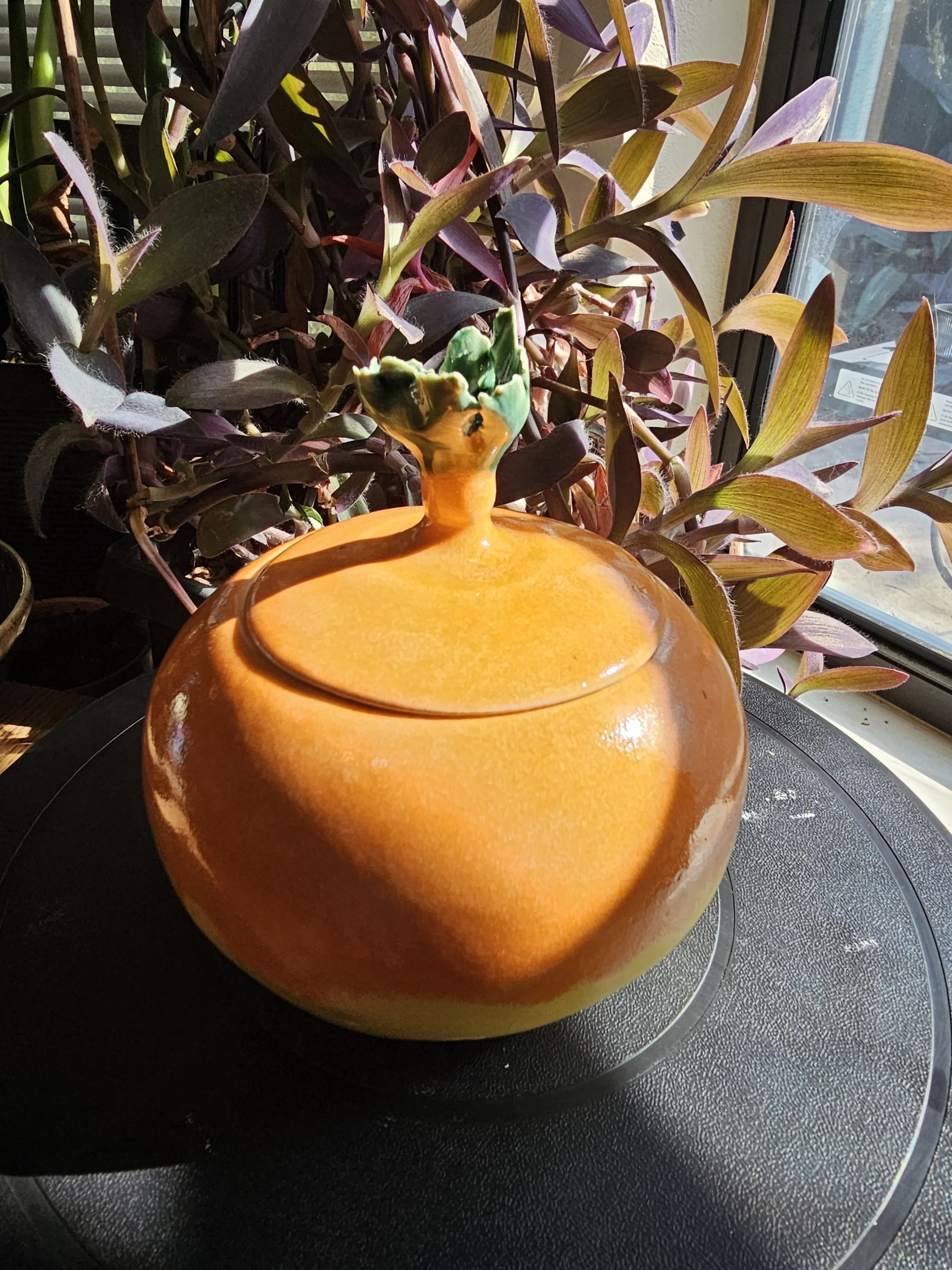 A small ceramic vase with a green and orange glaze, placed on a black surface near a window, with a plant with dark leaves in the background.
