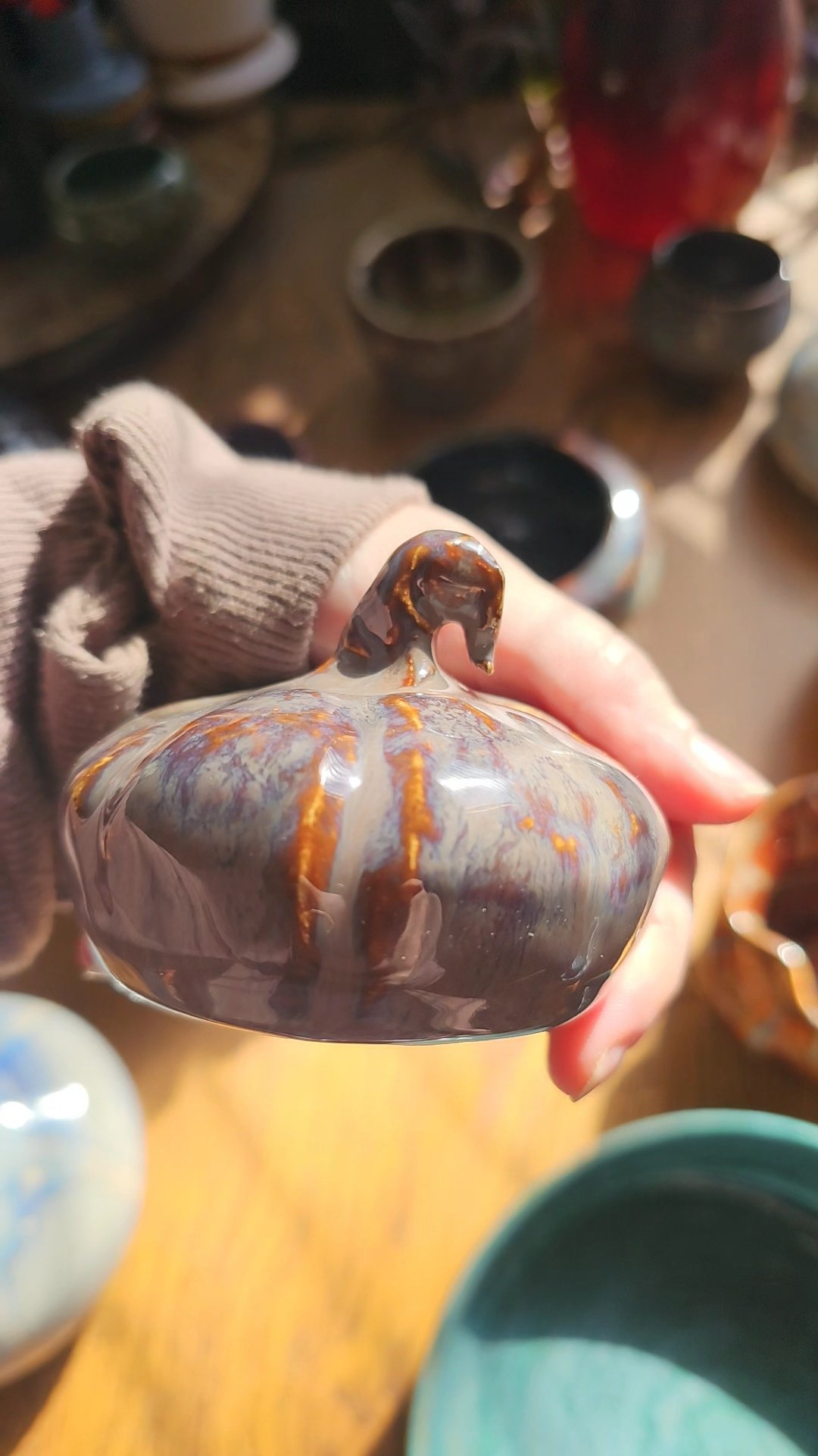 A person holding a small, round, glass object with a small horse-shaped handle, displaying a marbled pattern in brown, gray, and orange tones. The background shows shelves with various other glass items.