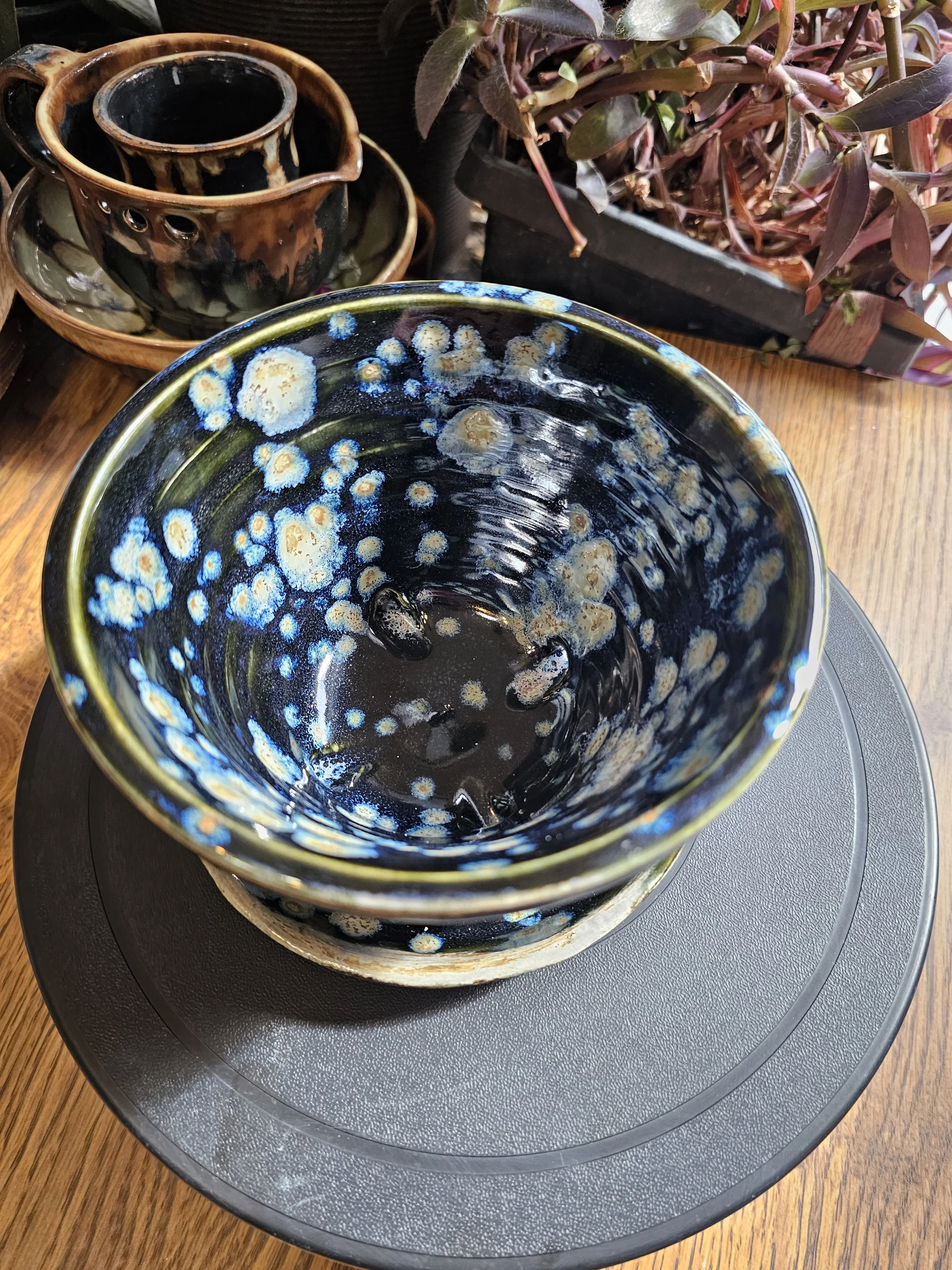 A black ceramic bowl with blue and beige accents, sitting on a black circular tray with a textured surface, placed on a wooden table. In the background, there are two matching ceramic cups and saucers with black and brown glaze, along with a black re