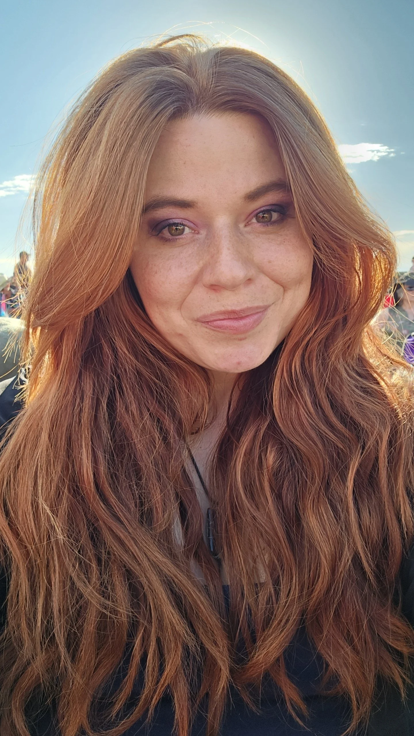 Close-up of a woman with long red hair, light skin, and freckles, smiling, outdoors in sunlight.