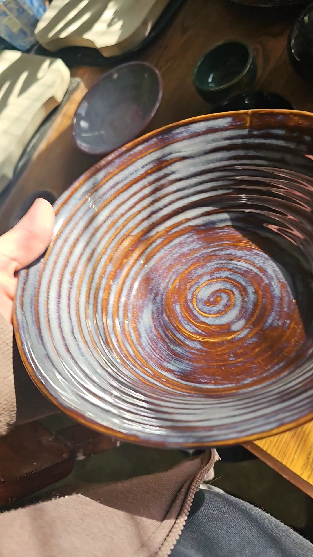A person holding a round ceramic plate with a spiral design in shades of orange, brown, and white. The plate has a glossy finish and is part of a set, with other similar plates visible on a wooden table in the background.