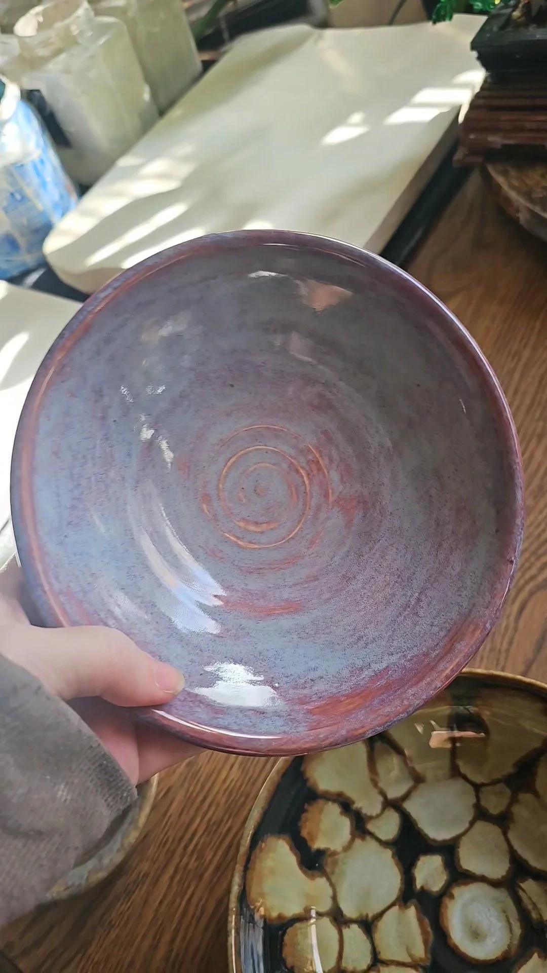 View of the bottom of a ceramic bowl with concentric circles, held by a person's hand, with various decorative ceramic items and wooden surfaces in the background.