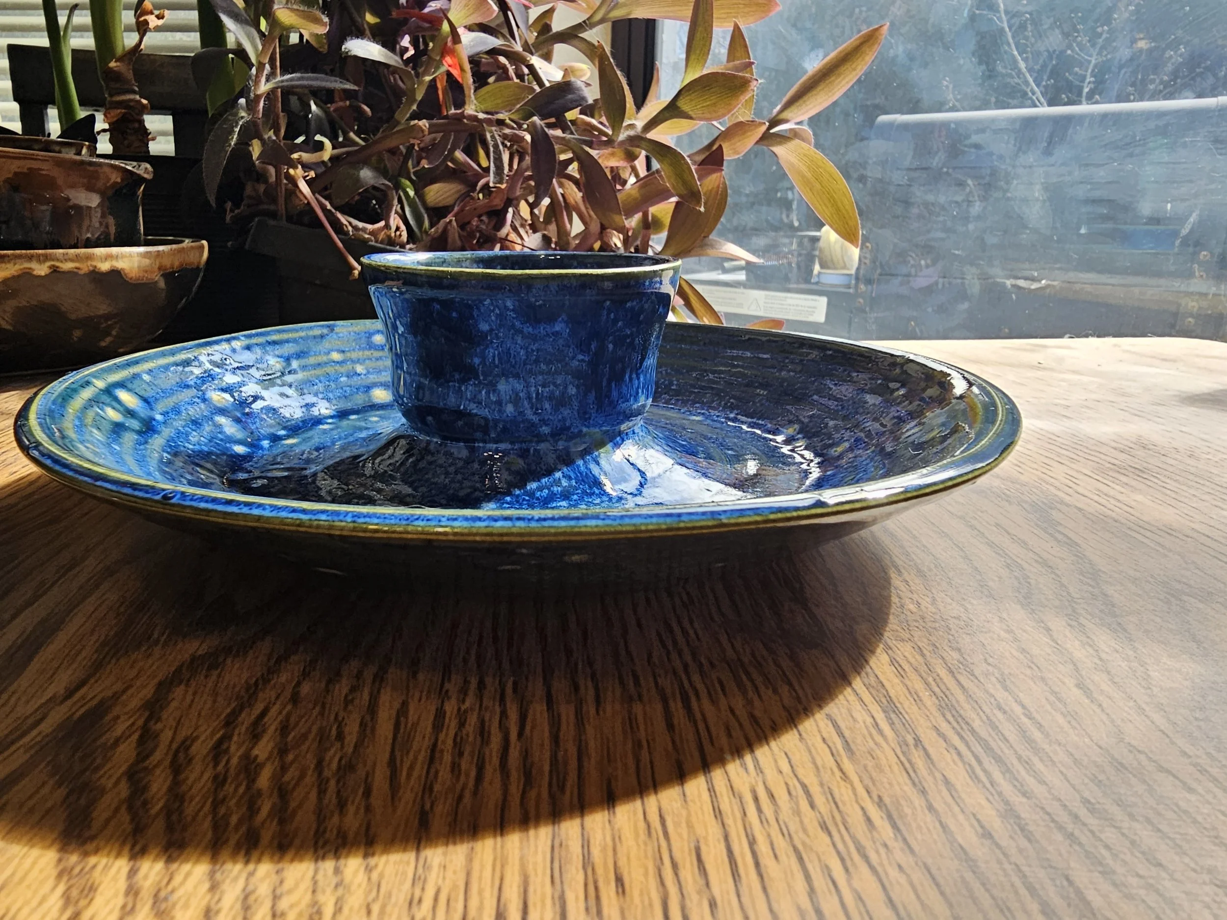 A blue ceramic chip and dip sitting on a matching blue ceramic plate on a wooden table, with potted plants on a windowsill in the background.
