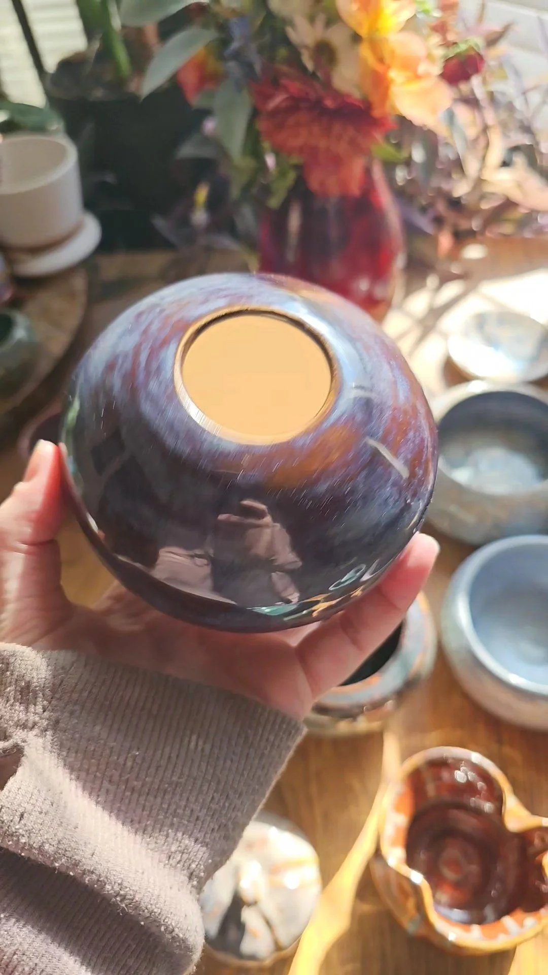 A person holding a dark glazed ceramic bowl, with other bowls and vases visible on a wooden surface in the background.